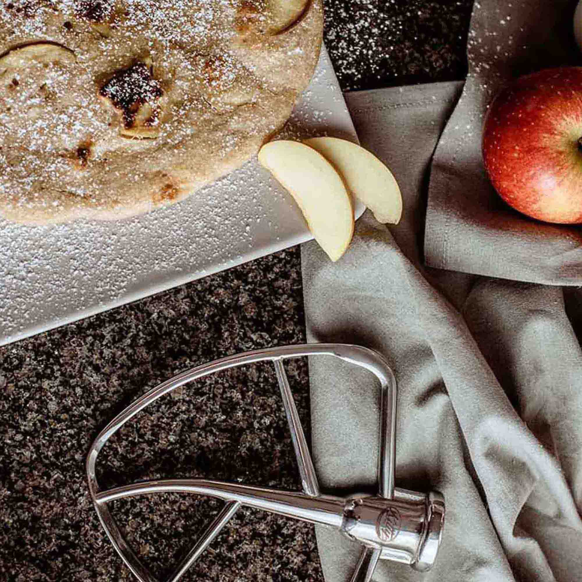 Ein Apfelkuchen auf einem weißen Teller, mit Apfelscheiben und einem roten Apfel. Ein Mixeraufsatz liegt daneben.