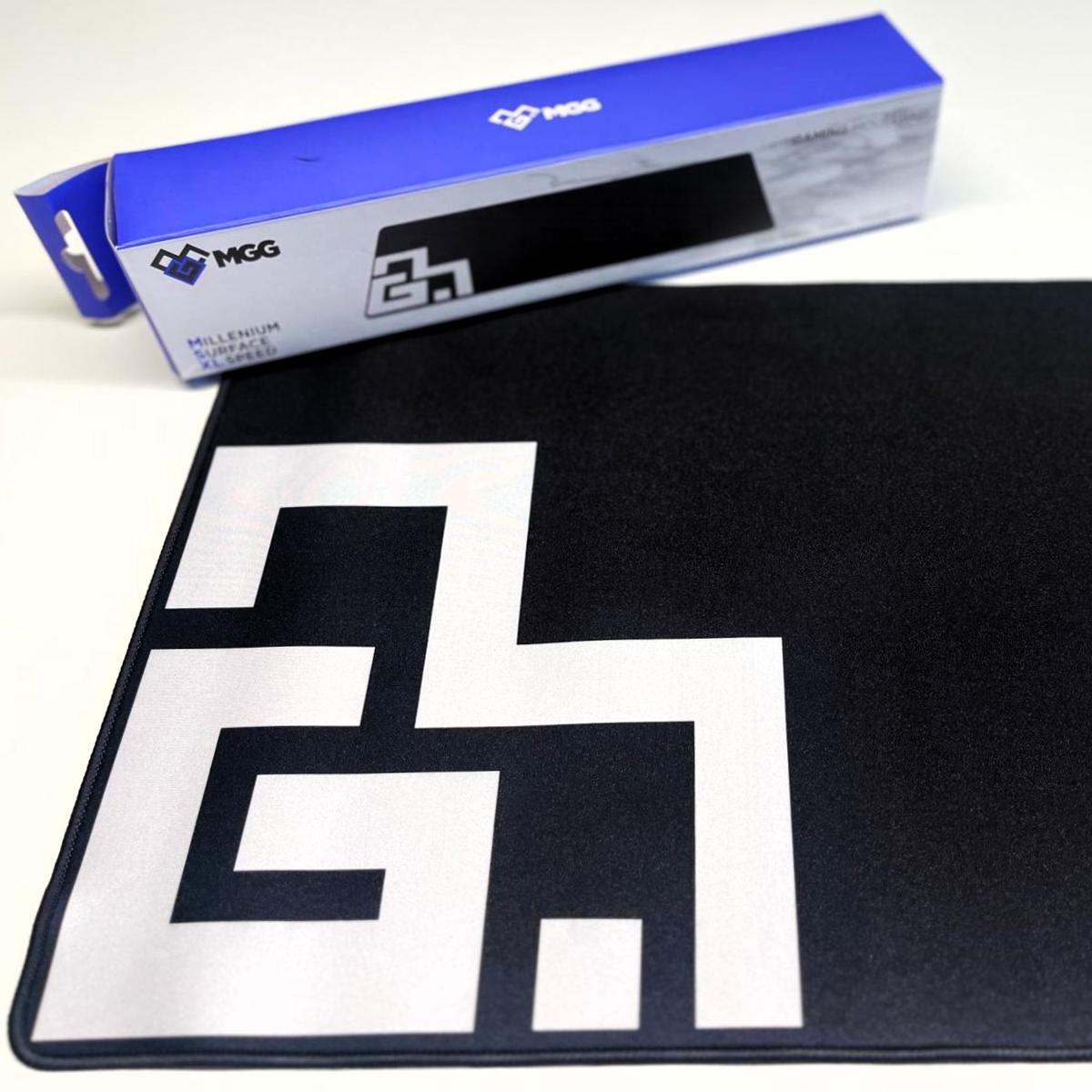 Alfombrilla de ratón gaming negra con logo blanco y embalaje de fondo.
