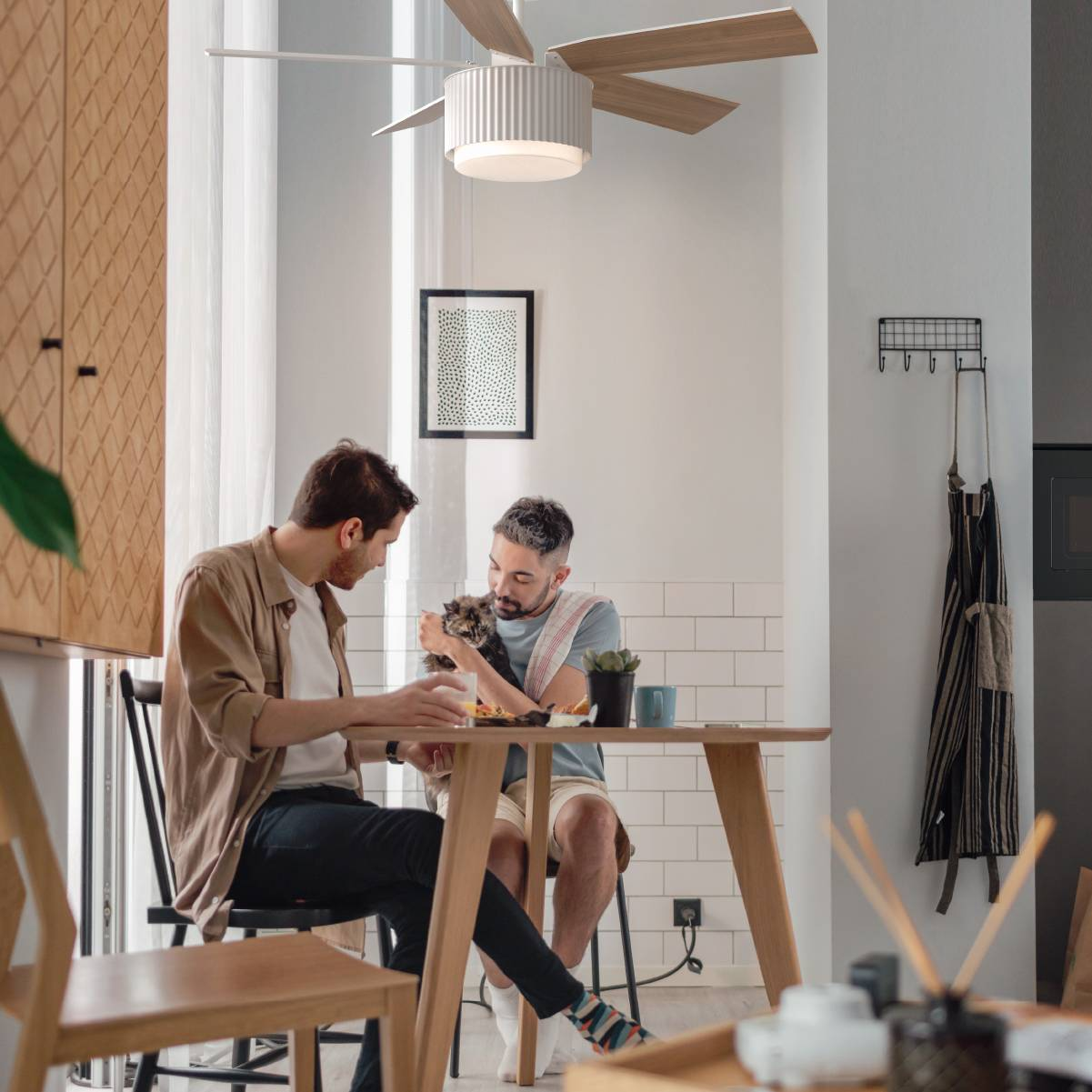 Dos hombres, uno con un gato, en una mesa. Ventilador de techo visible arriba.