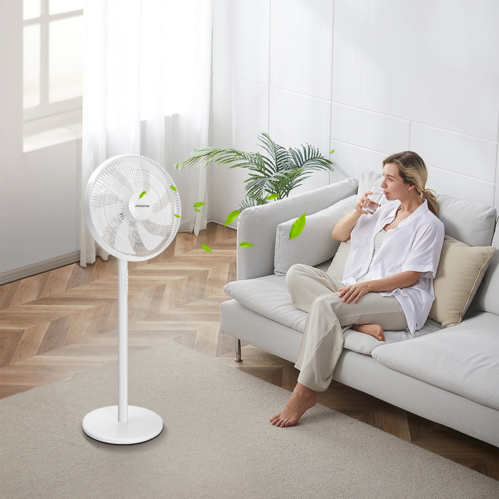 Mujer bebe agua cerca de un ventilador blanco. Hojas verdes vuelan. Un sofá y ventana están detrás.