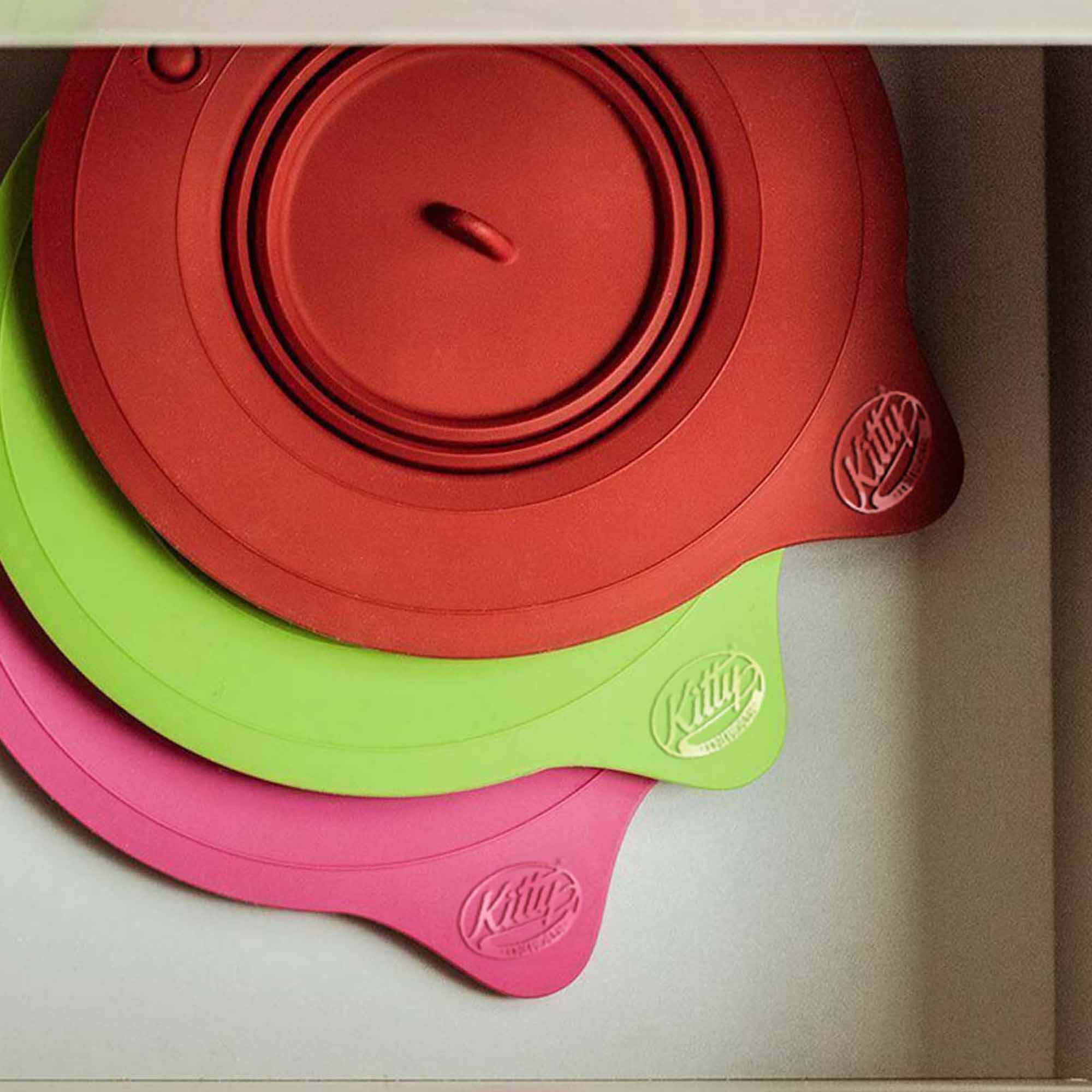 Drei bunte Silikondeckel in einer Schublade gestapelt, rot, grün und rosa, mit einem Markenlogo auf jedem.
