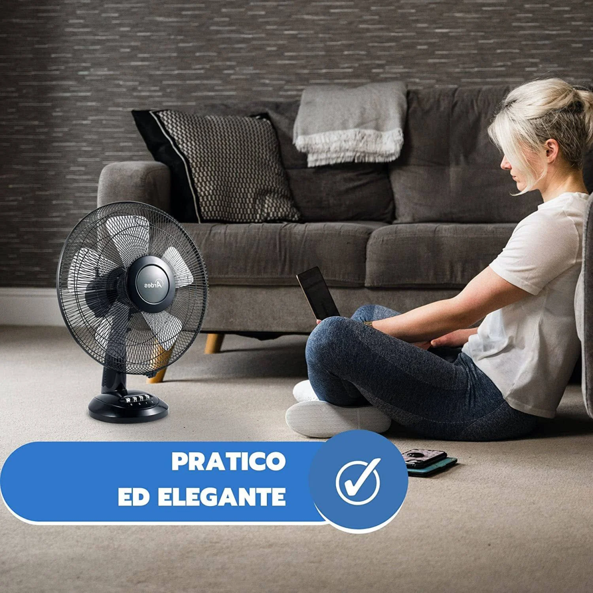 Kobieta siedzi na podłodze obok sofy, używając laptopa. Czarny wentylator na pierwszym planie.