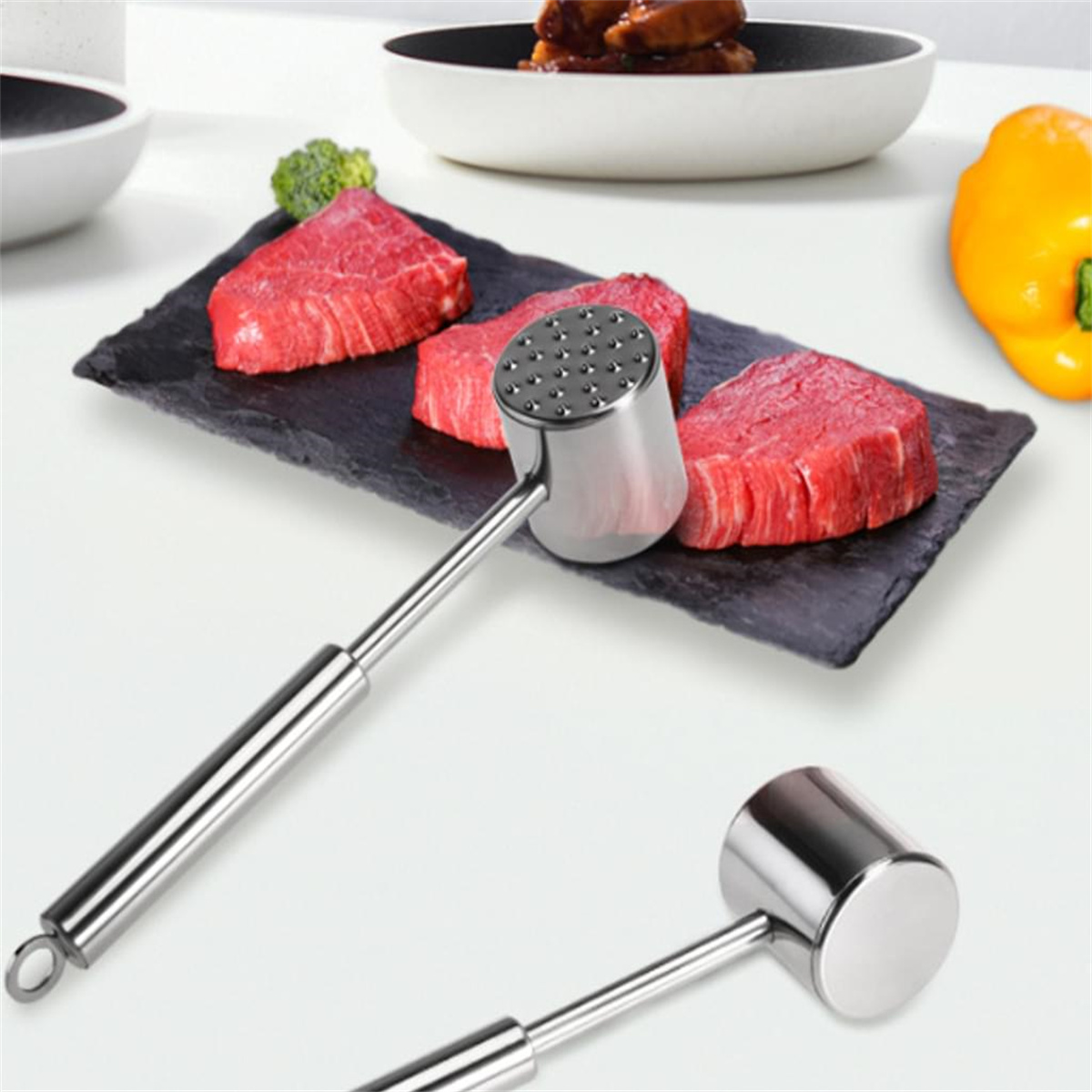 Fleischklopfer und rohes Fleisch auf einem Schieferbrett mit anderen Lebensmitteln wie Brokkoli und gelbem Paprika.