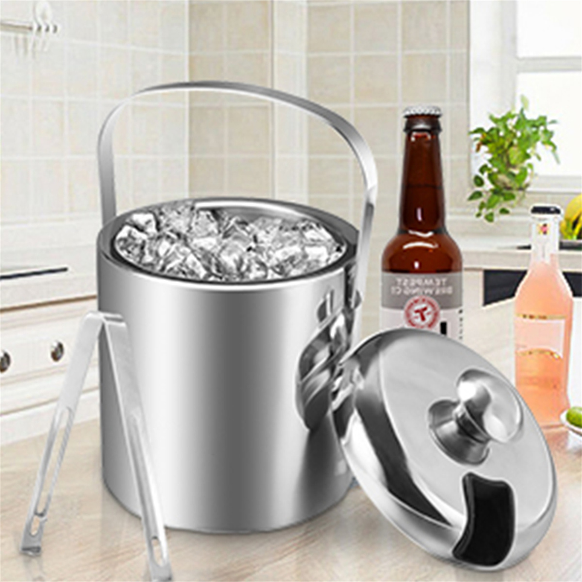 Ein Edelstahl-Eisbehälter mit Eis und Zangen, neben einer Bierflasche und einem rosa Getränk.