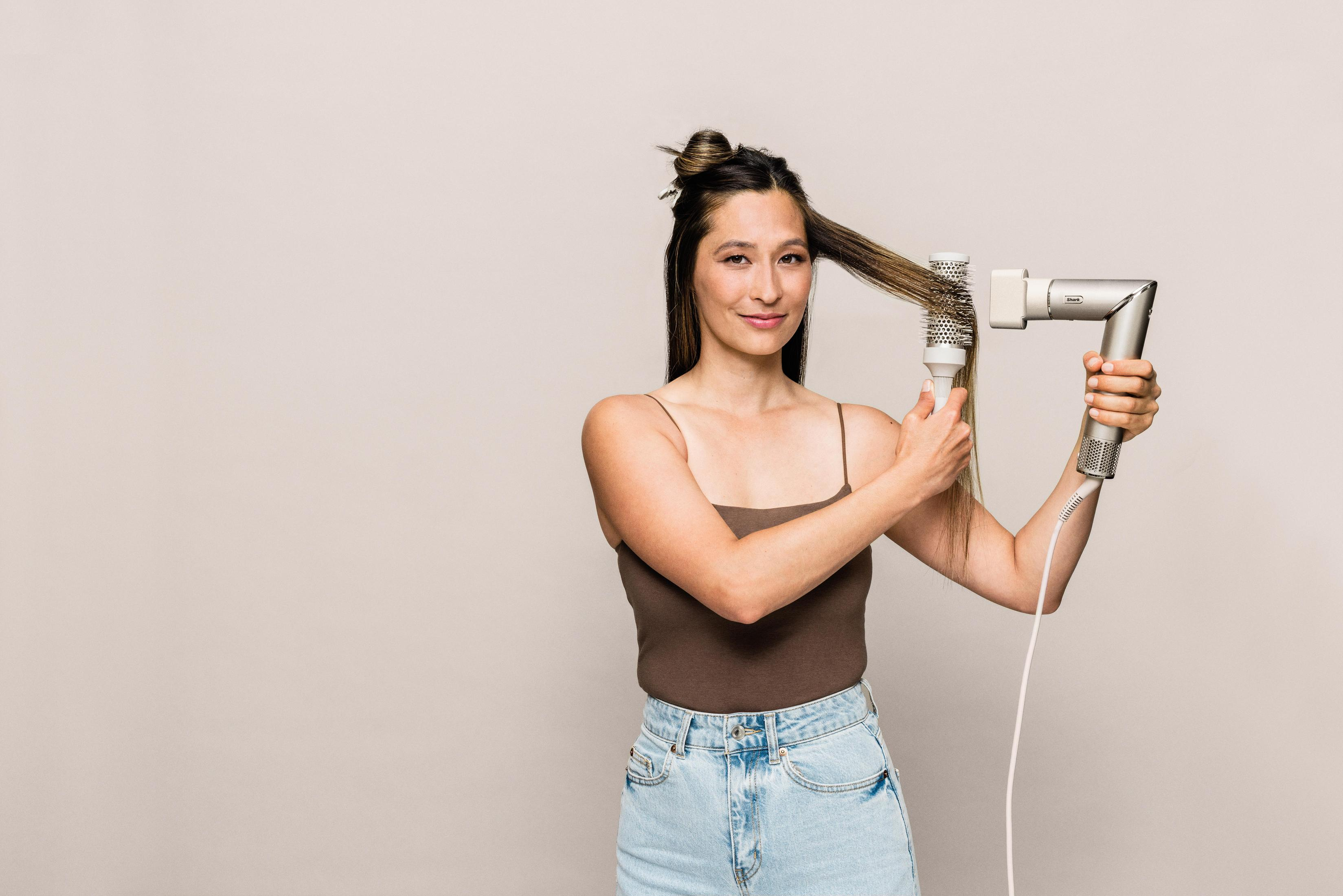 Una donna sta usando un asciugacapelli e una spazzola rotonda sui suoi capelli. Sta sorridendo.