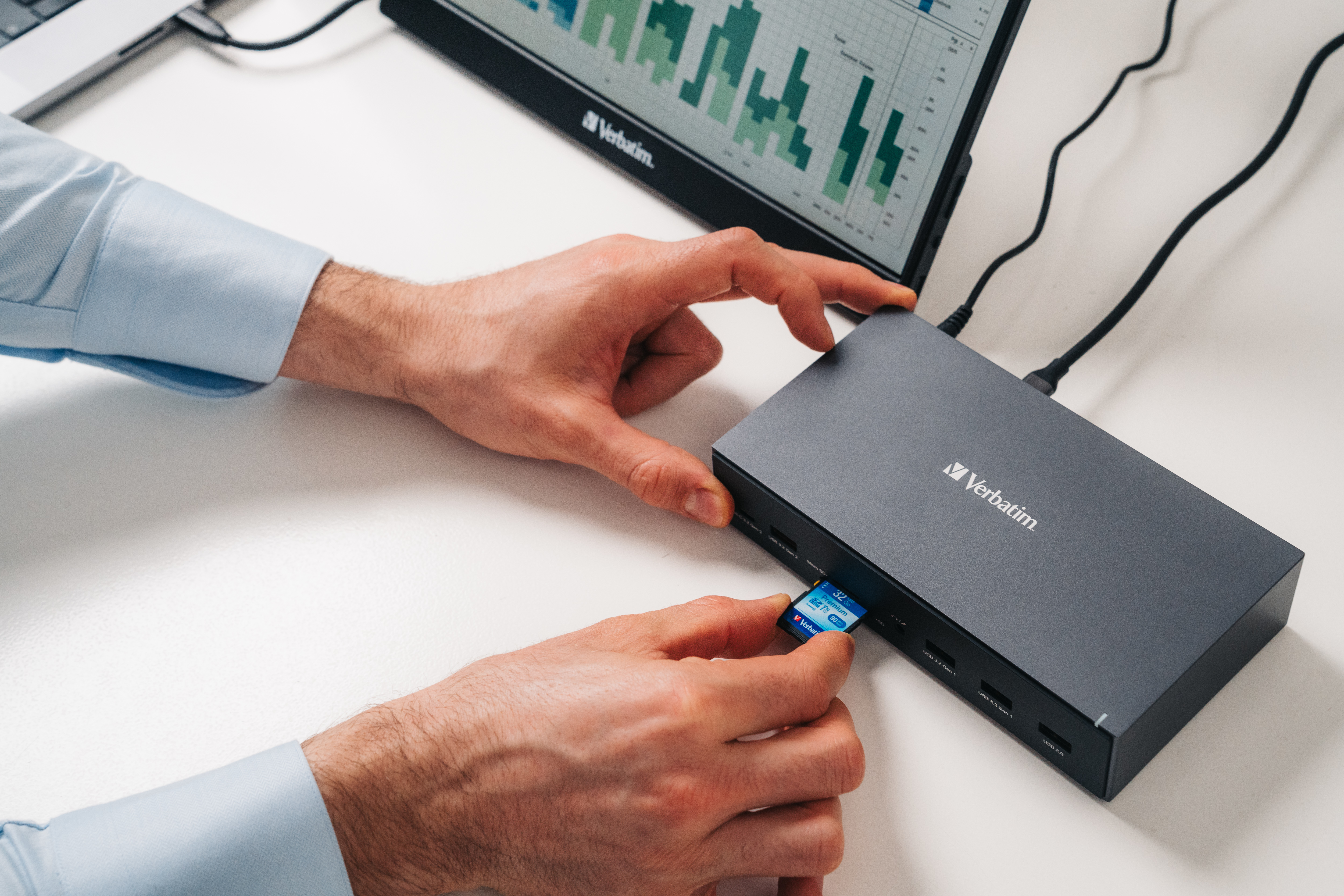 Hände stecken eine blaue SD-Karte in einen schwarzen Verbatim-Hub ein, ein Laptop und ein Monitor stehen auf dem weißen Schreibtisch.