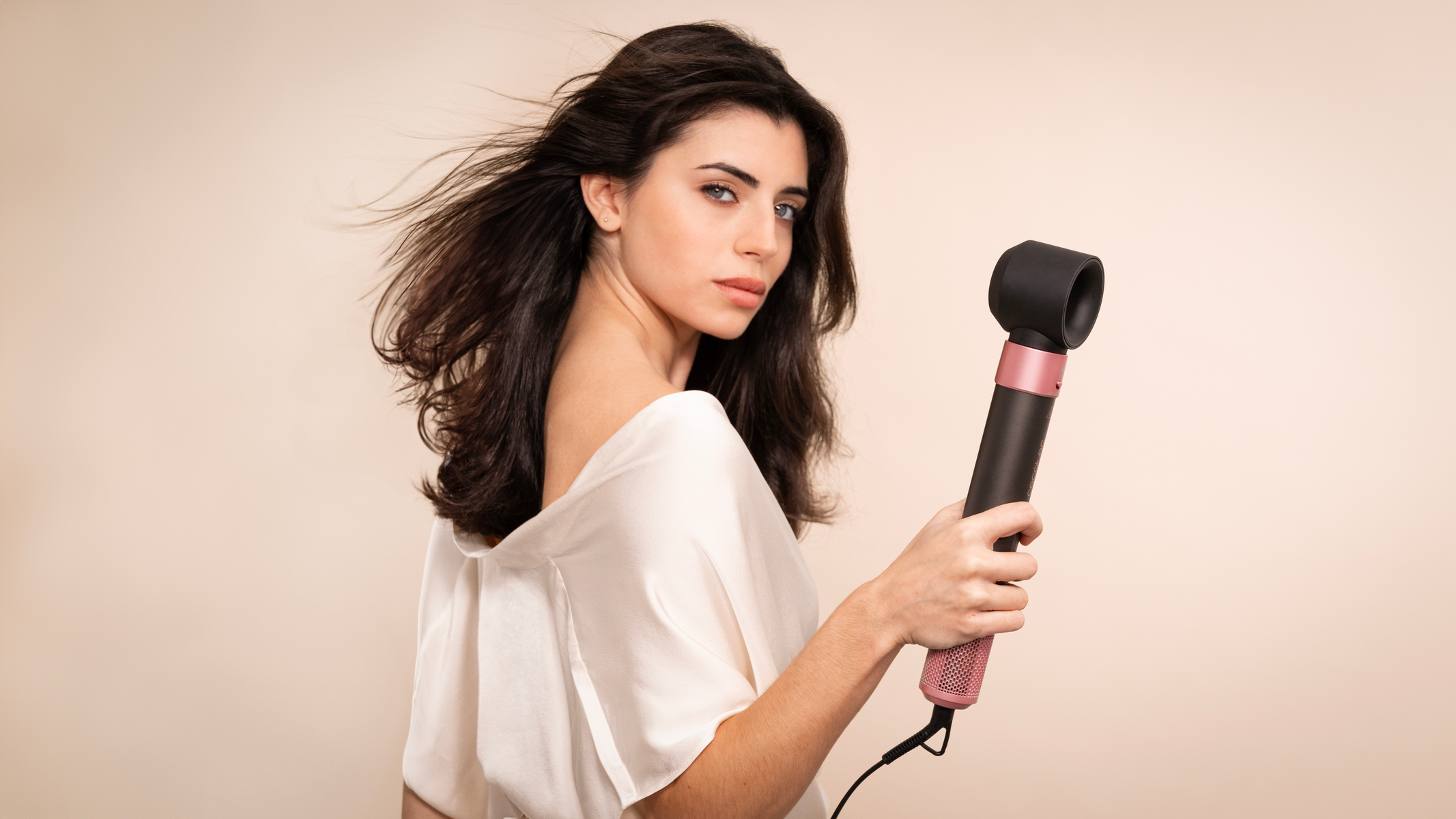 Vrouw die een haardroger vasthoudt. Ze heeft bruin haar en draagt een witte top. De achtergrond is beige.
