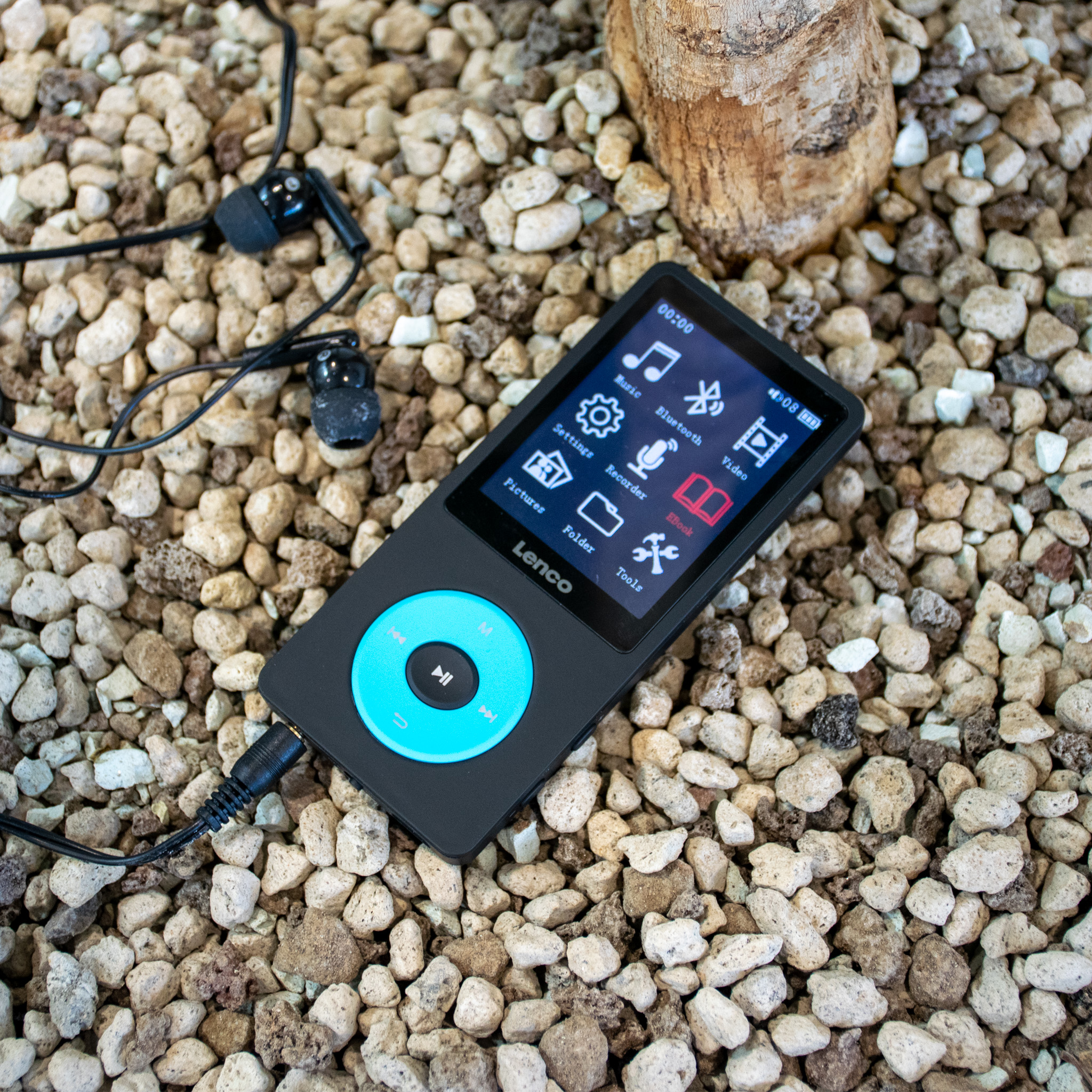 Ein schwarzer Lenco MP3-Player mit einem Bildschirm, der Musikbedienelemente anzeigt, auf einem Kiesbett.
