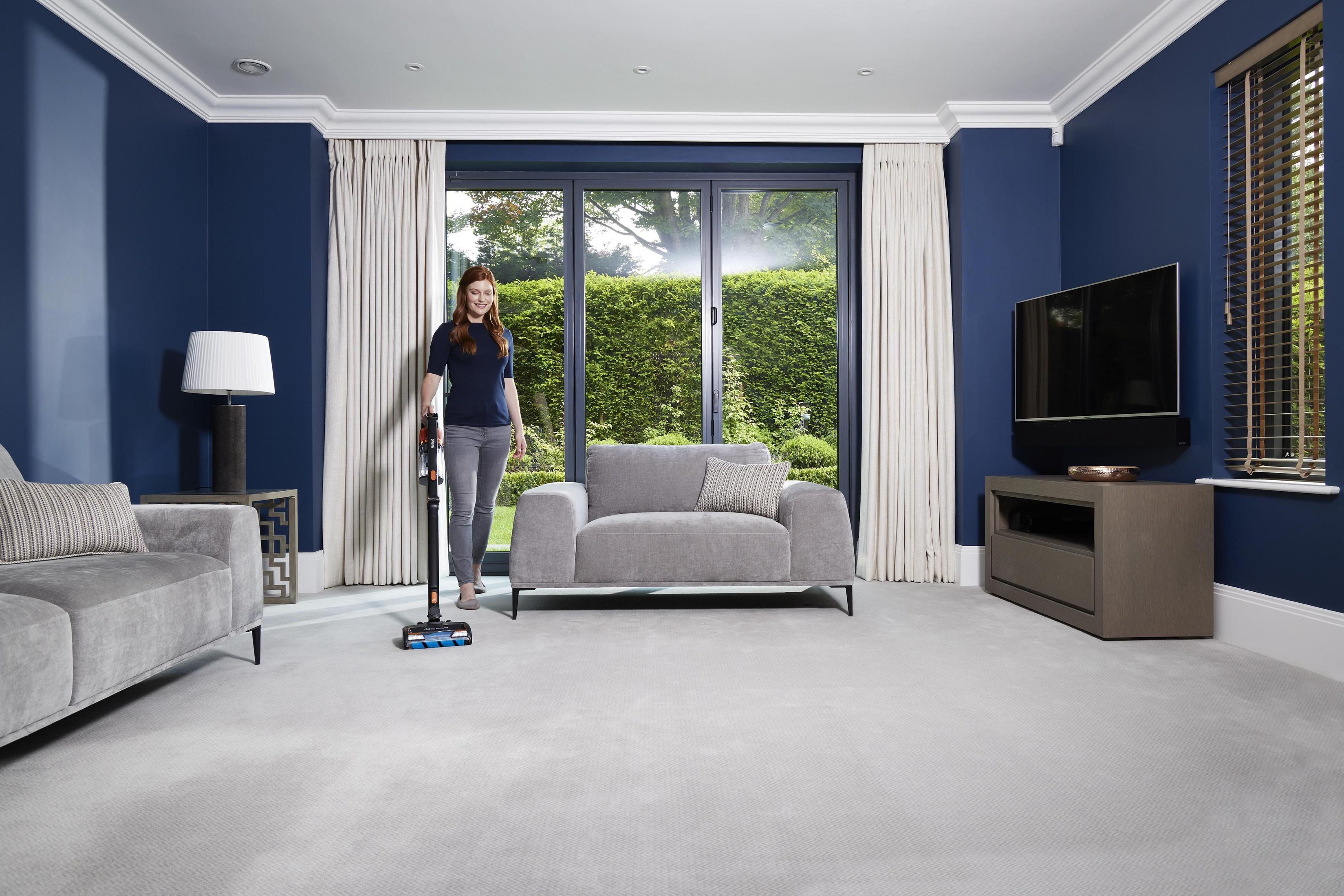 Una mujer aspirando una habitación. La habitación tiene paredes azules, un sofá gris y un televisor.