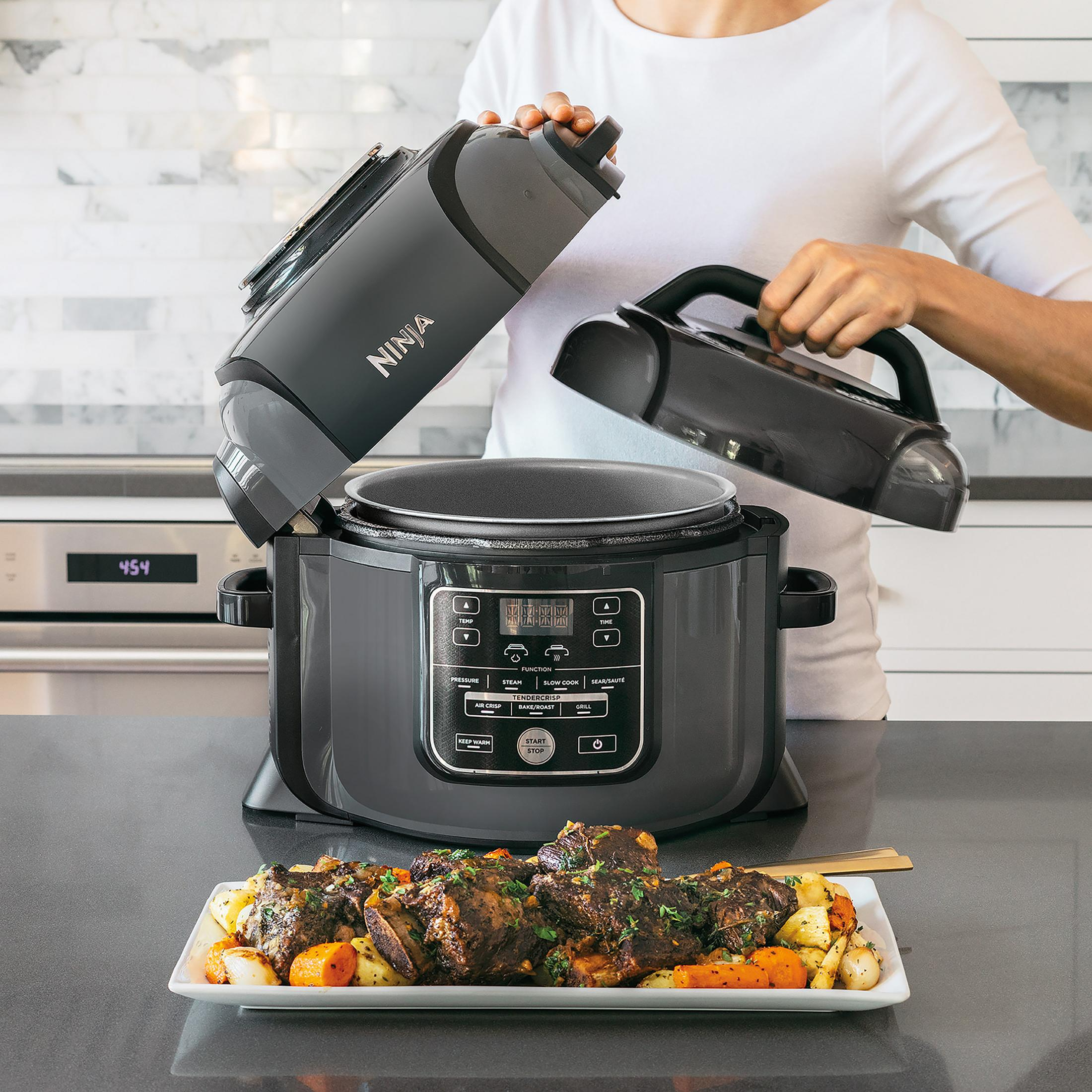 Una persona apre una pentola multifunzione Ninja, rivelando un piatto di carne e verdure cotte.