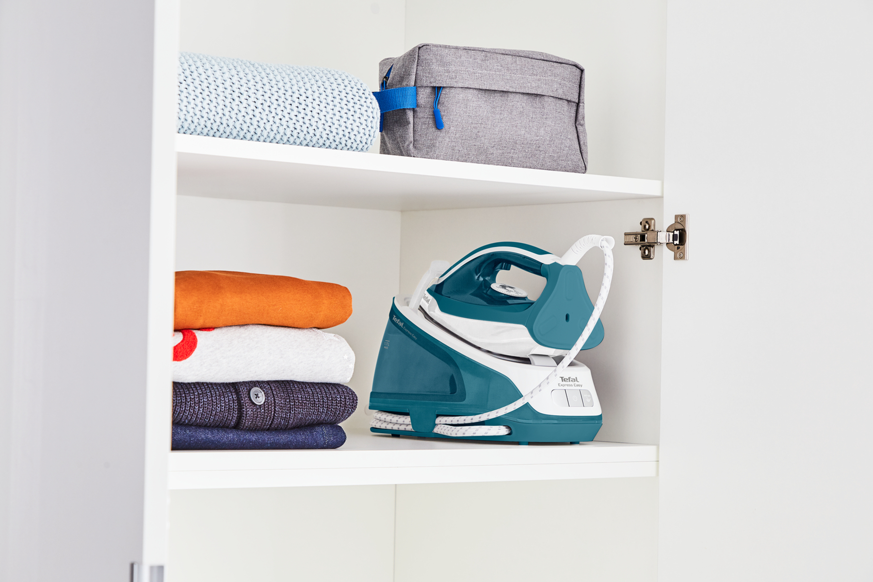 Bügeleisen und gefaltete Kleidung in einem weißen Schrank. Eine graue Reisetasche steht oben.