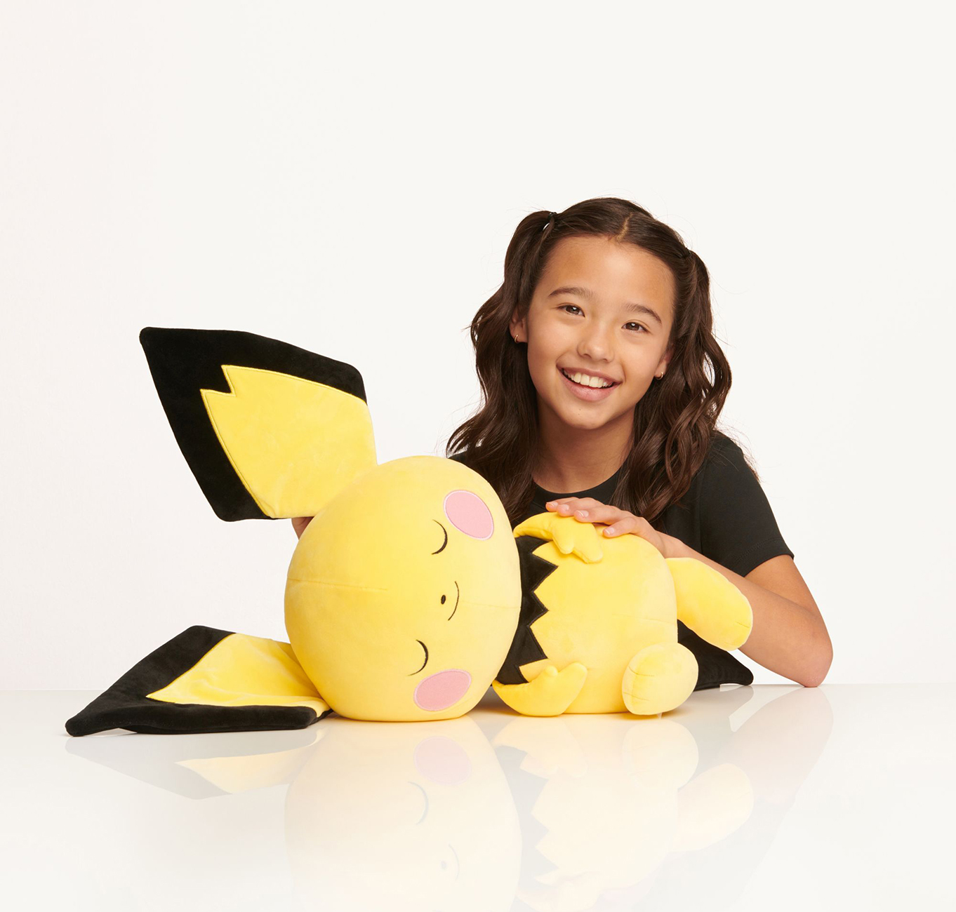 Ein lächelndes Mädchen, das ein gelbes Pikachu-Plüschtier auf einer weißen Oberfläche hält.