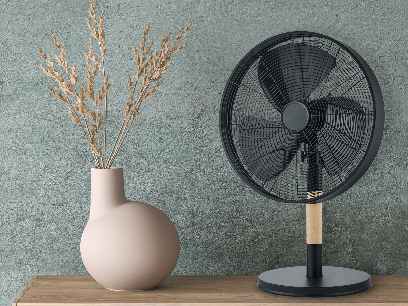 Ein schwarzer Standventilator mit Holzbein und eine Vase mit getrockneten Pflanzen auf einem Holztisch.