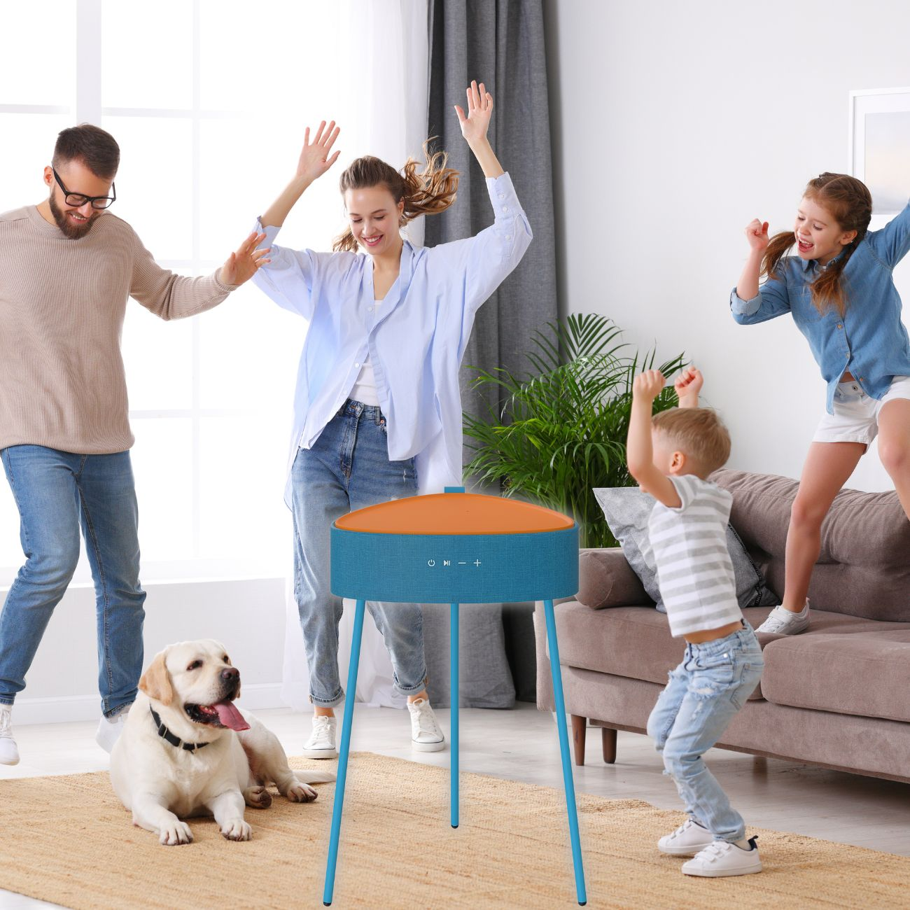 Familie tanzt um einen blauen Lautsprecher mit orangefarbenem Deckel. Ein Hund liegt auf dem Teppich. Fröhliche Szene.