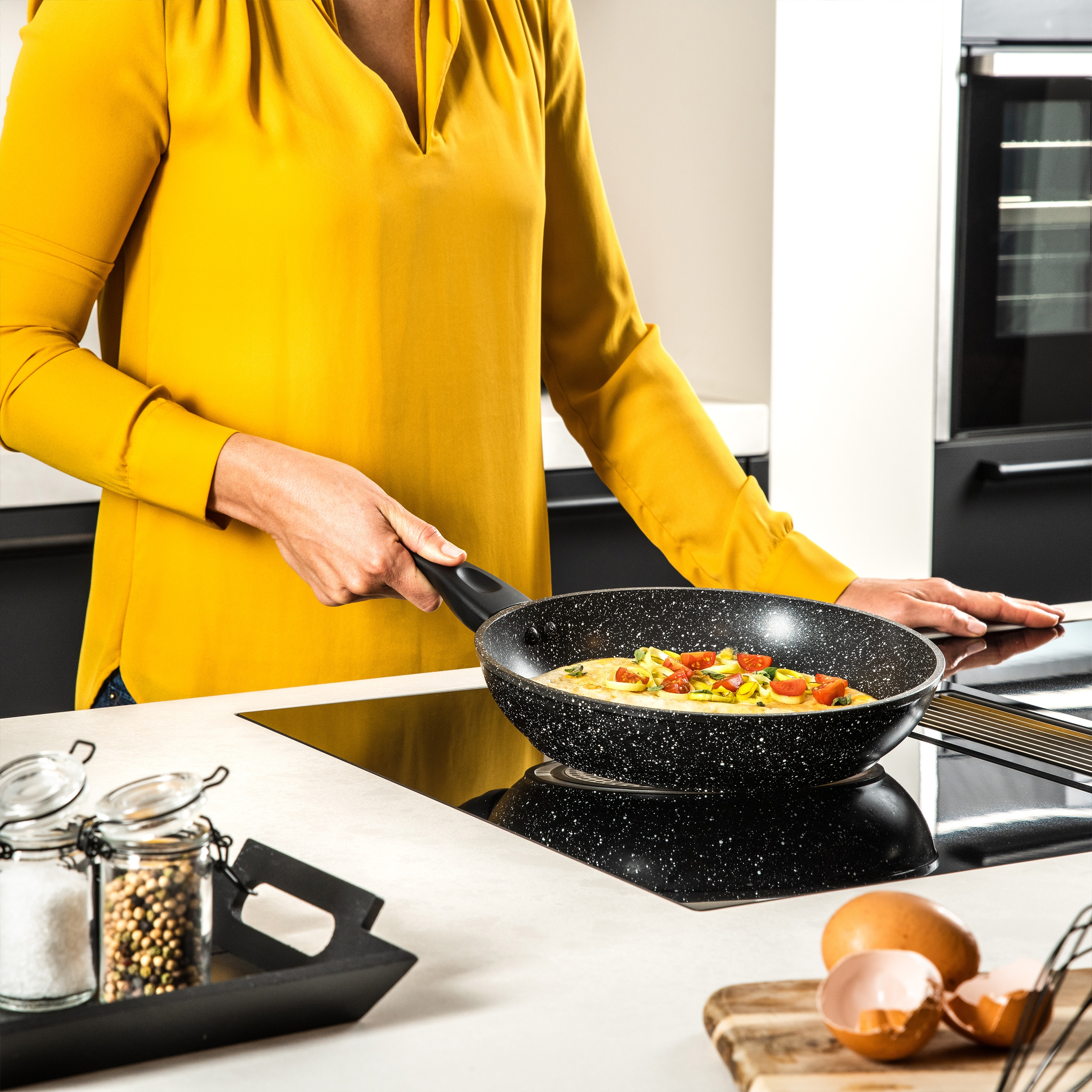 Eine Person hält eine Pfanne mit gekochtem Essen. Zutaten, Gläser und Herd im Hintergrund.