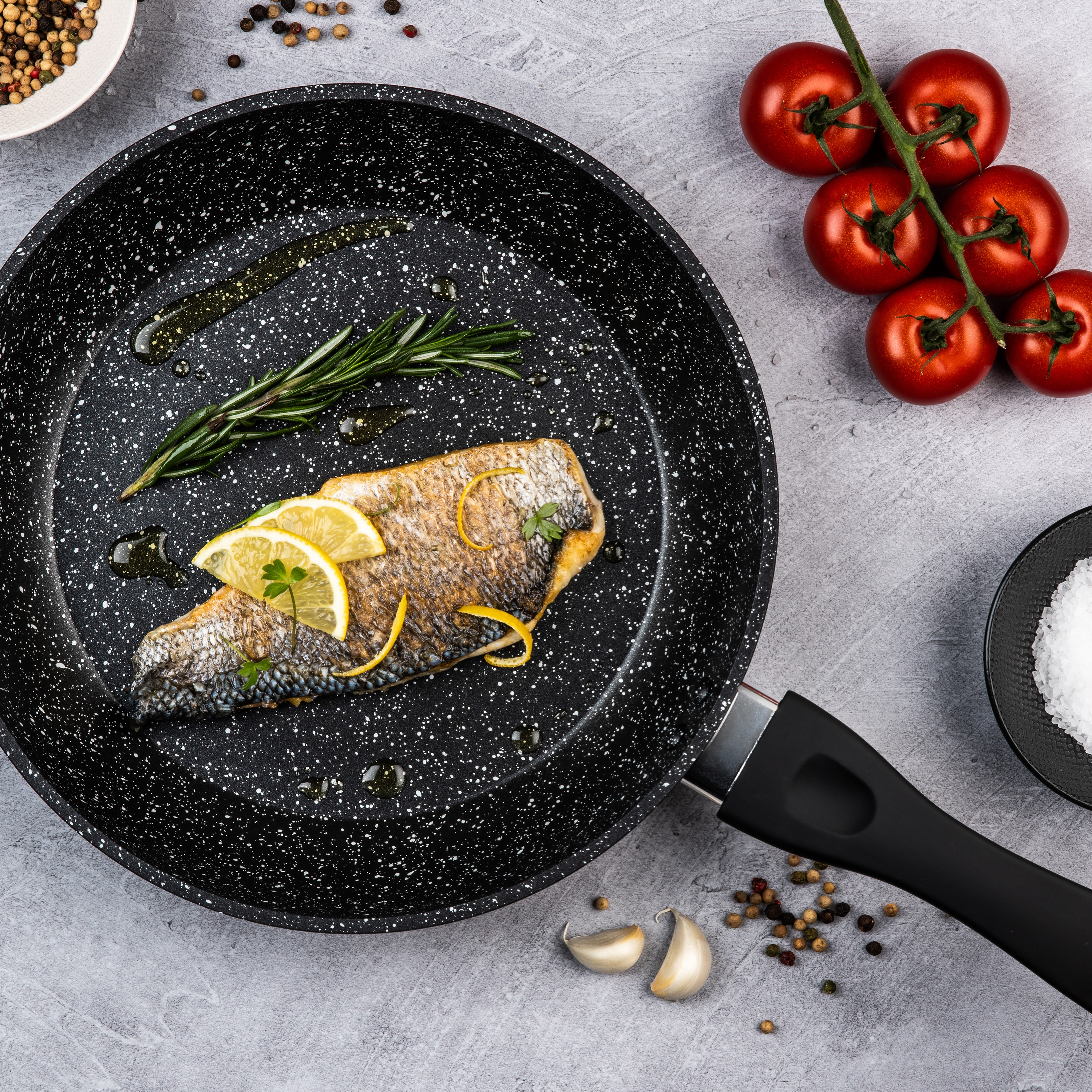 Eine Bratpfanne mit gekochtem Fisch, Zitronenscheiben, Rosmarin und Tomaten. Die Pfanne ist gesprenkelt.