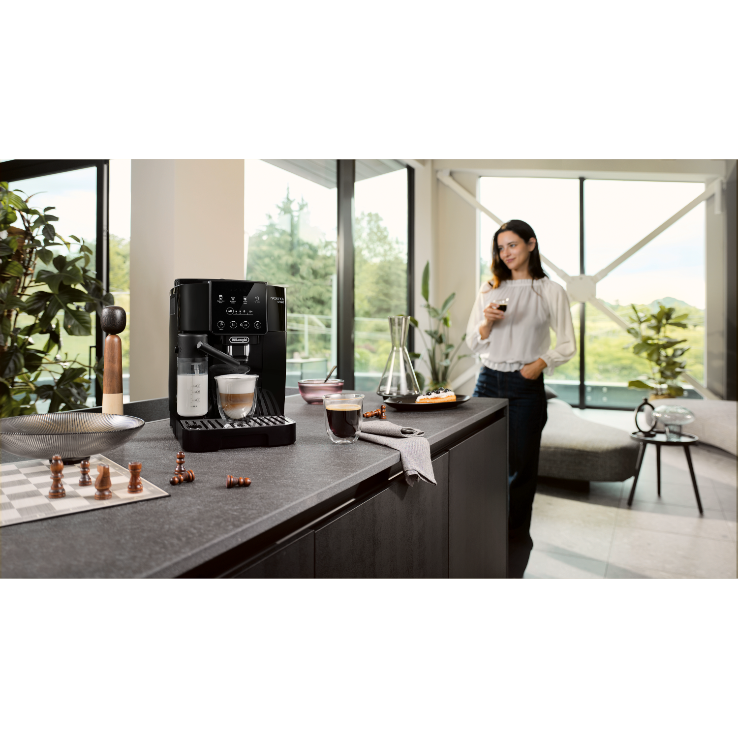 Kobieta przy ekspresie do kawy z szachami i osobą w tle trzymającą szklankę.