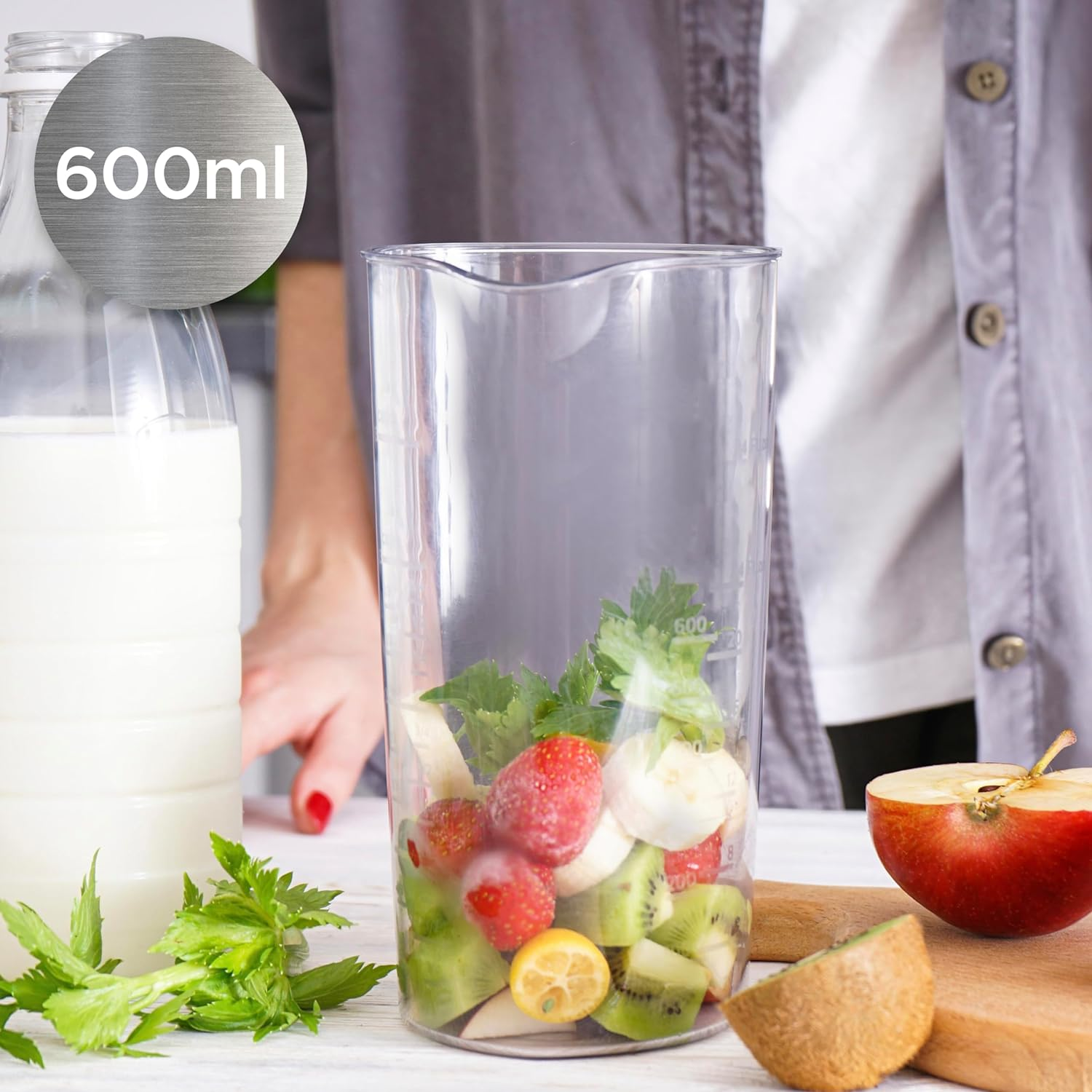 Ein Mixerbehälter ist mit Obst und Gemüse gefüllt. Eine Milchflasche und ein Apfel sind in der Nähe.