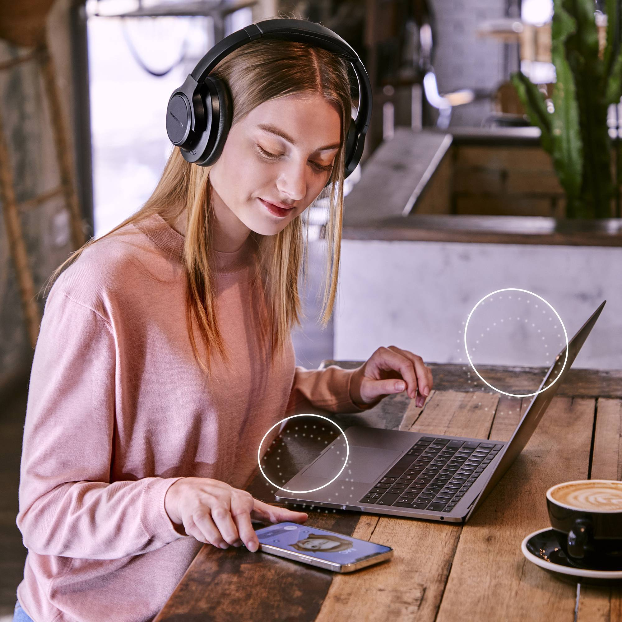 Donna al caffè con laptop e telefono. Cuffie indossate. Tazza di caffè sul tavolo. Tavolo in legno, sfondo chiaro.