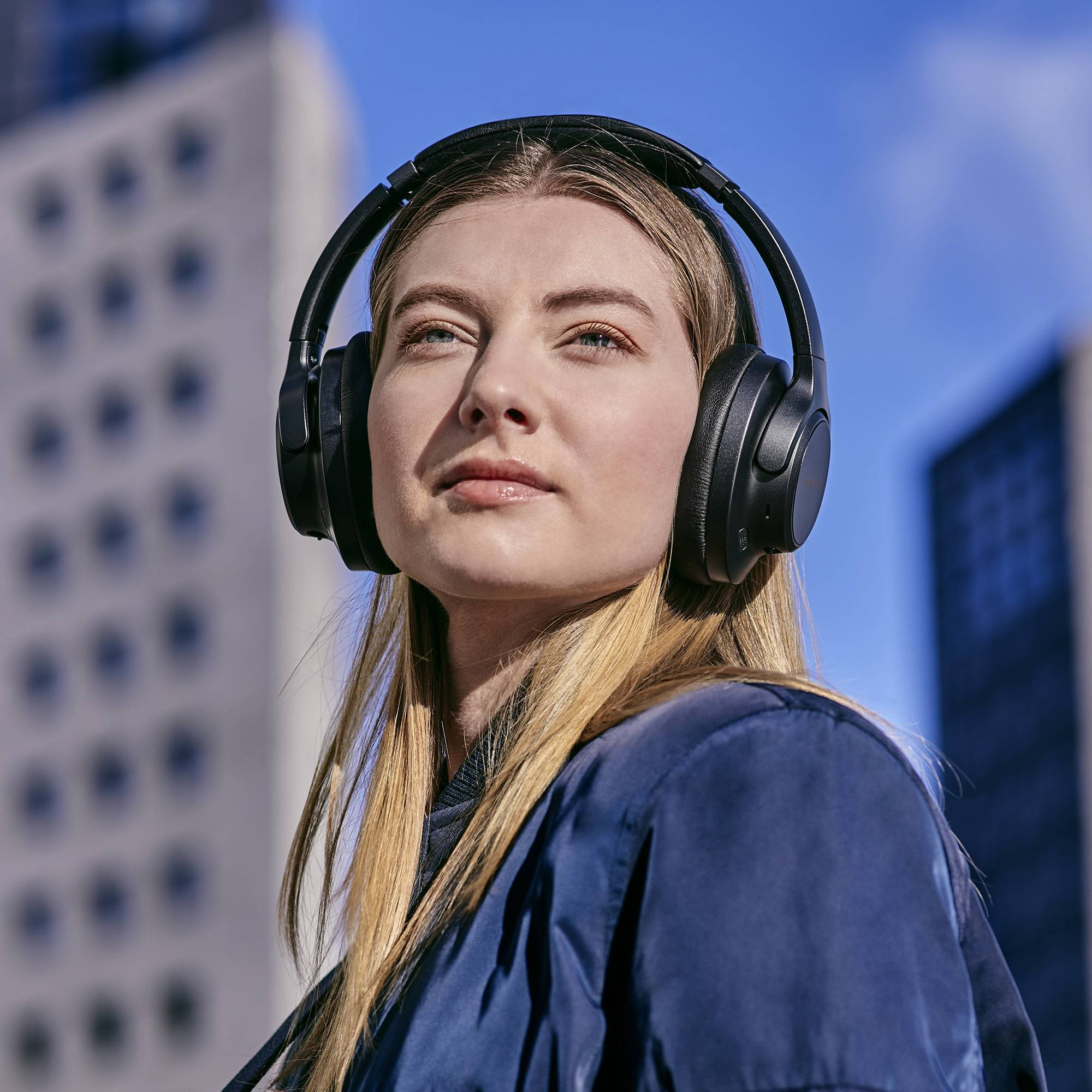 Una donna con i capelli biondi indossa cuffie nere, con uno sfondo di edifici urbani.