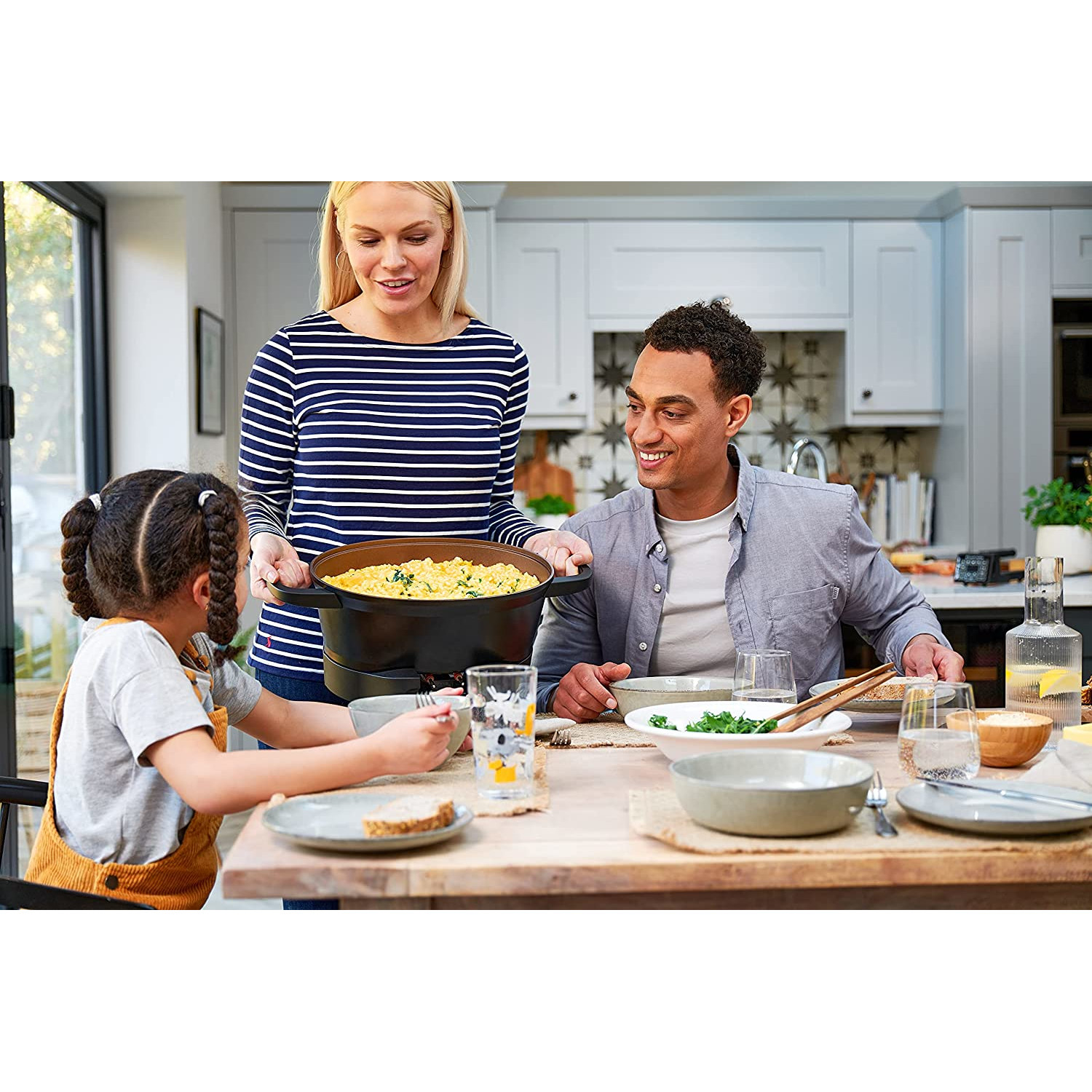 Familie am Tisch beim Essen. Frau serviert Essen aus einem schwarzen Topf. Helle Küche.
