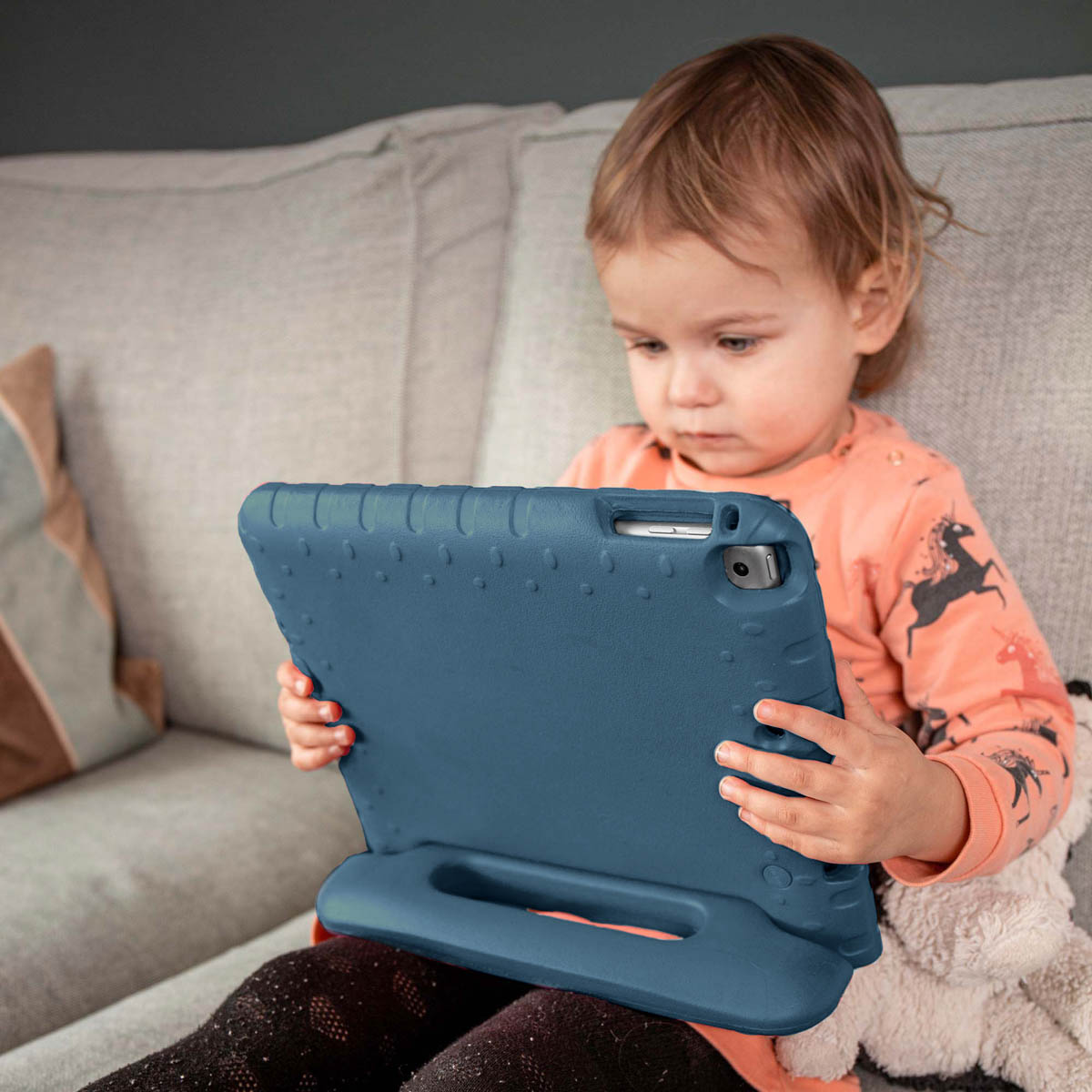 Een jong kind met een perzikshirt houdt een blauwe tablet voor een bank vast.