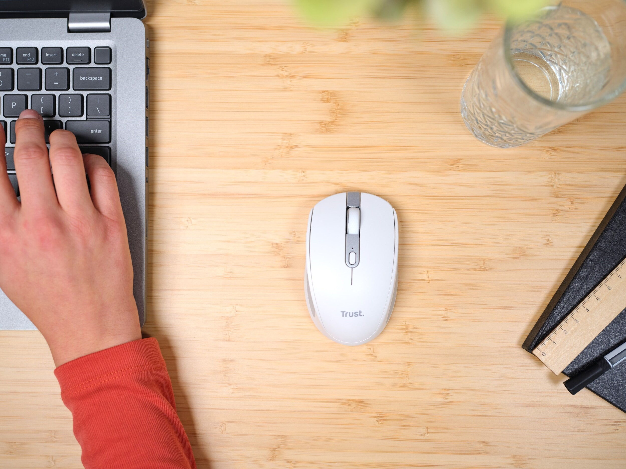 A person's hands using a laptop and a white mouse on a wooden table with a glass.