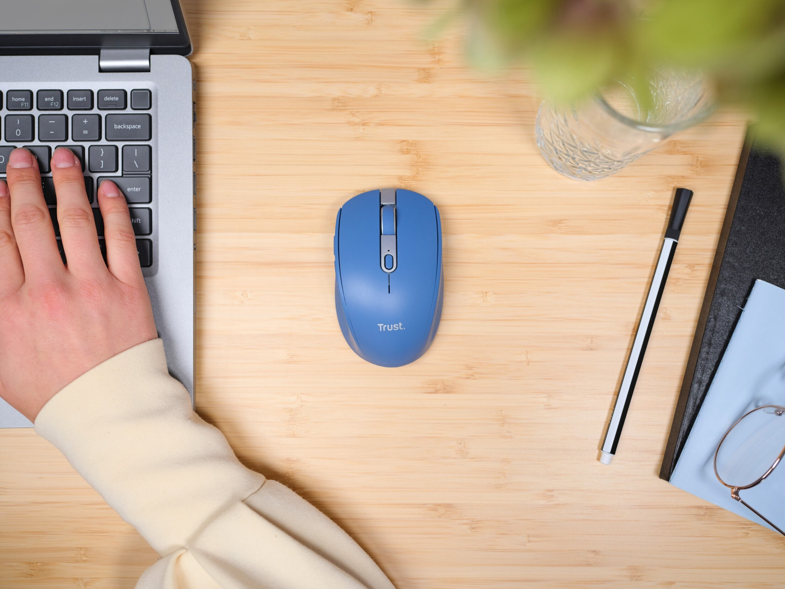 Dłoń na klawiaturze laptopa, niebieska mysz, długopis i szklanka na drewnianym stole.