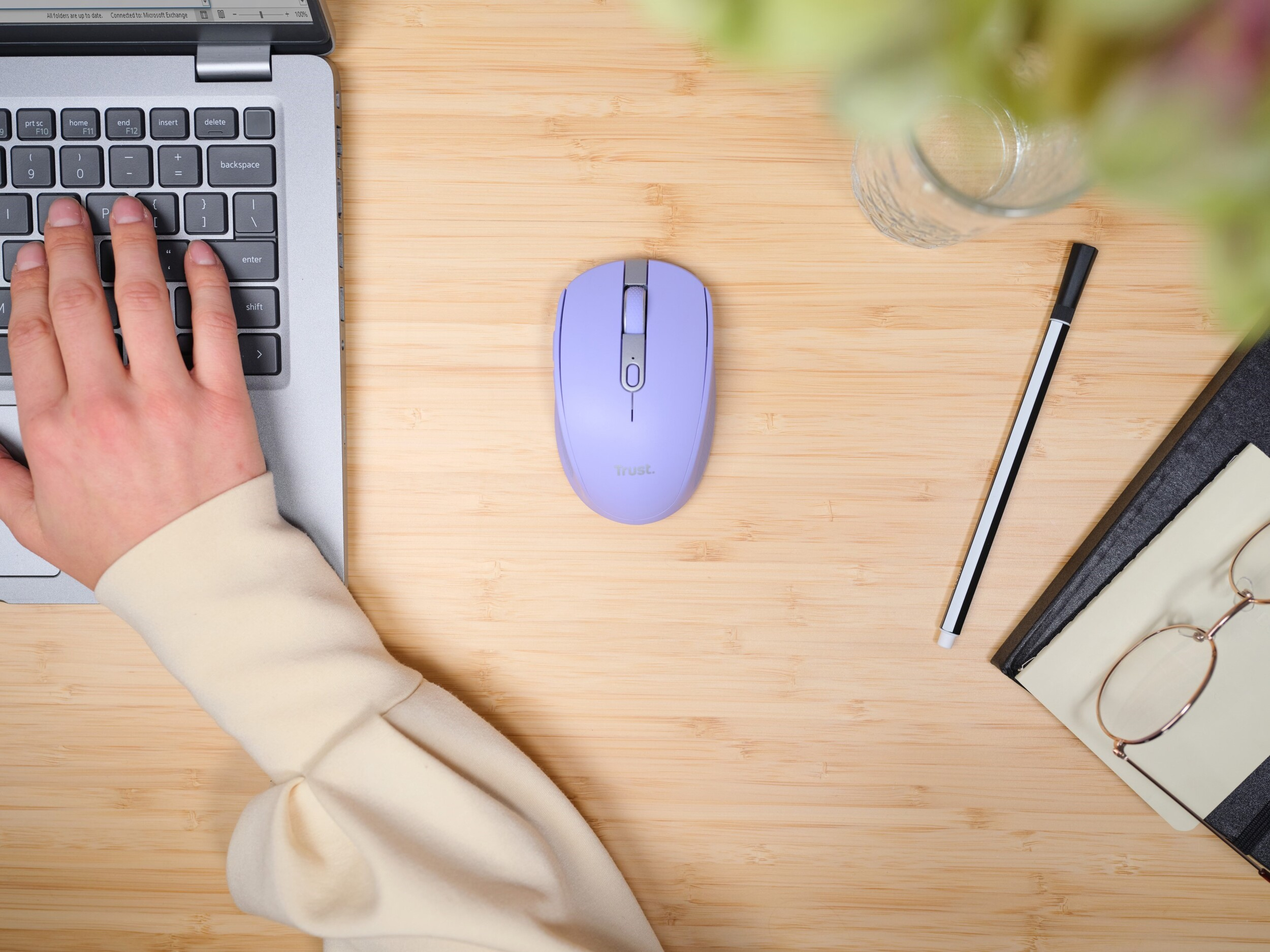 Laptop-Tastatur und eine lila Maus auf einem Holzschreibtisch mit Hand, Stift, Glas und Notizblock.