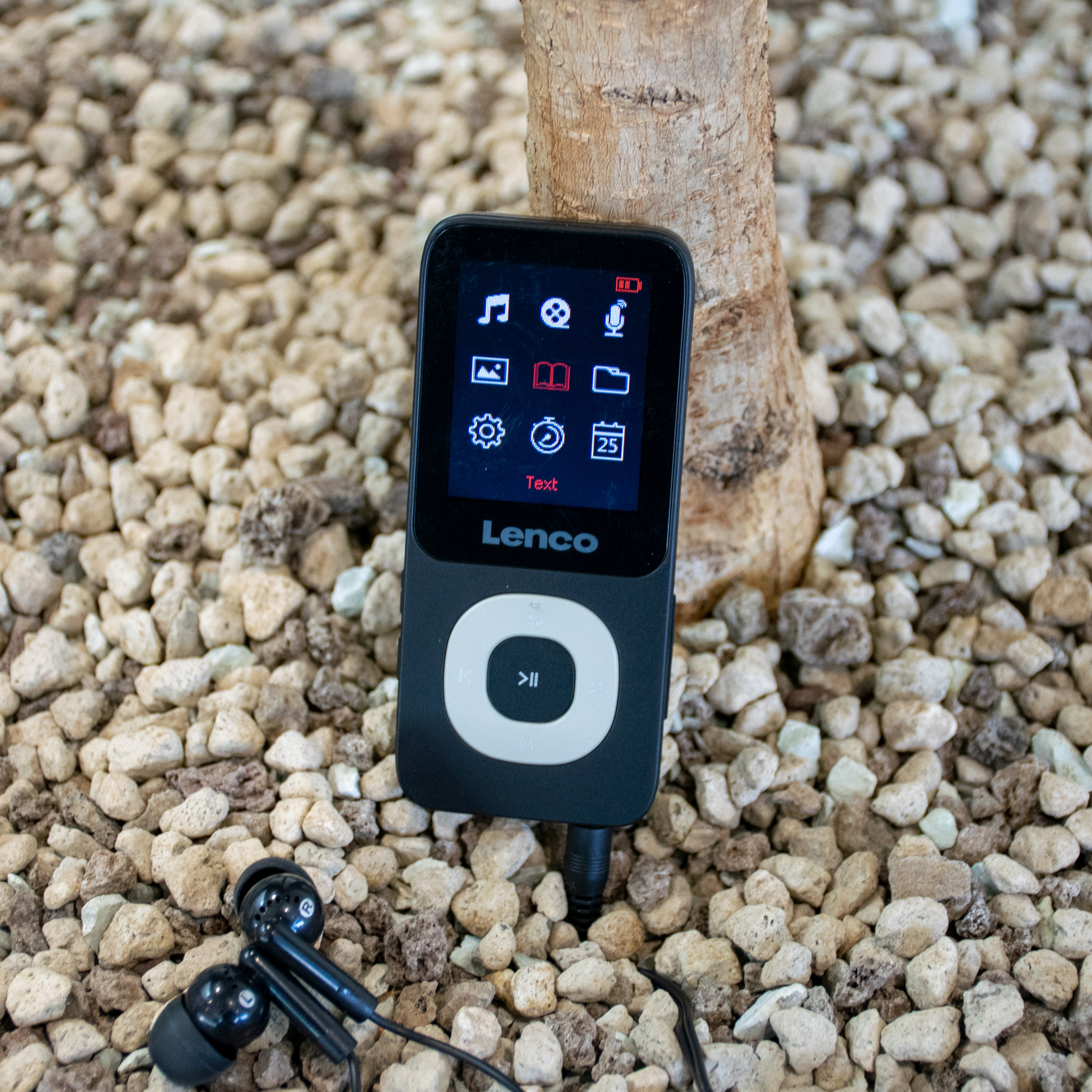 Lettore MP3 nero con schermo, cuffie e un tronco d'albero sullo sfondo.
