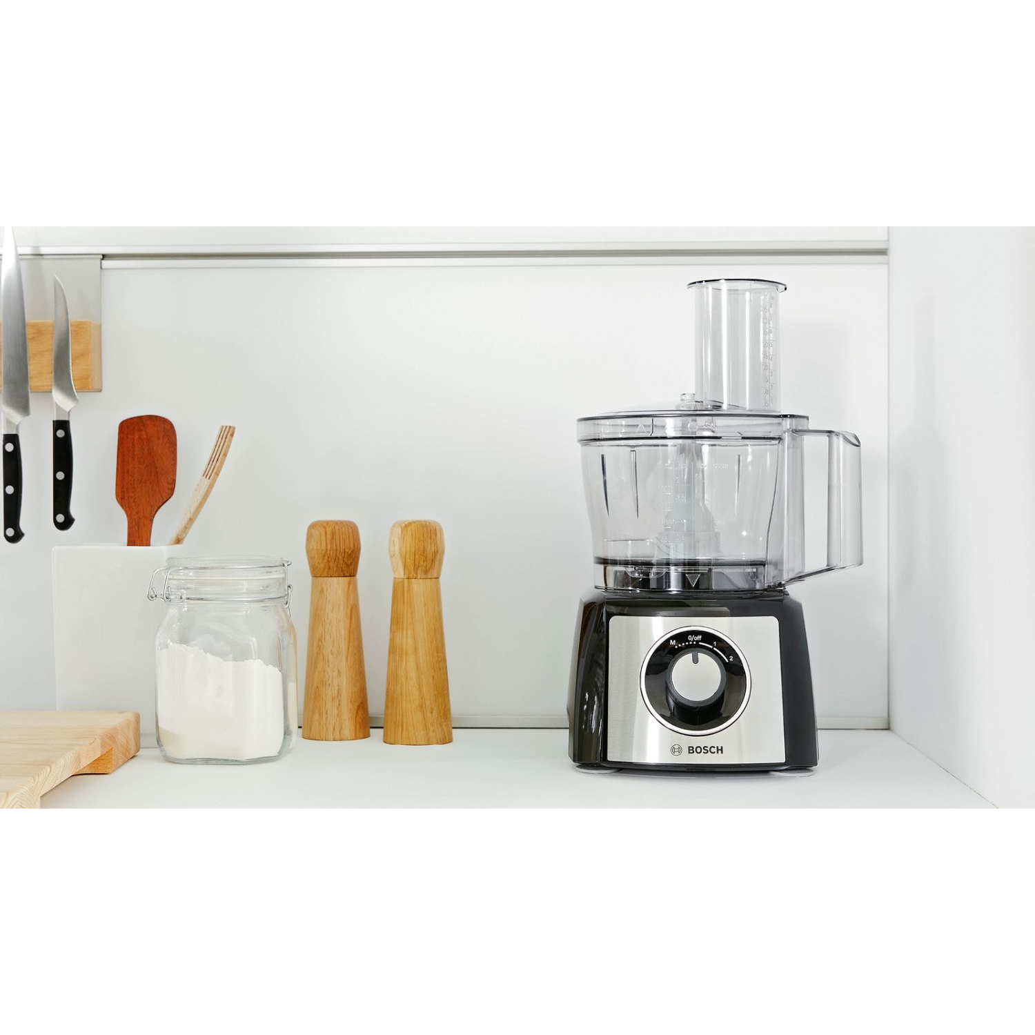 Robot kuchenny ze składnikami, nożami i drewnianymi przedmiotami na białej półce.