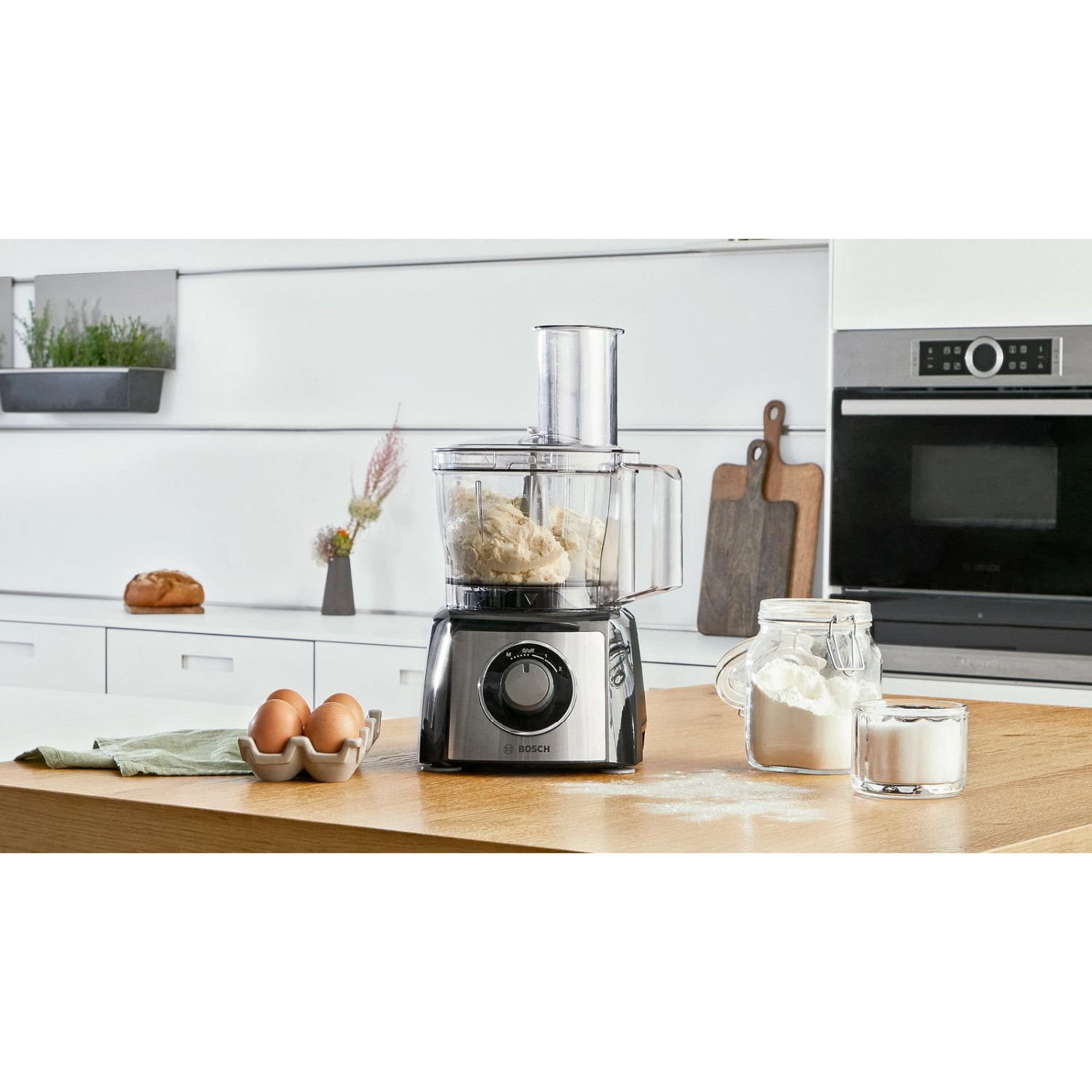 Robot kuchenny z ciastem, jajkami, mąką w słoiku i deską do krojenia, wszystko na drewnianej powierzchni.