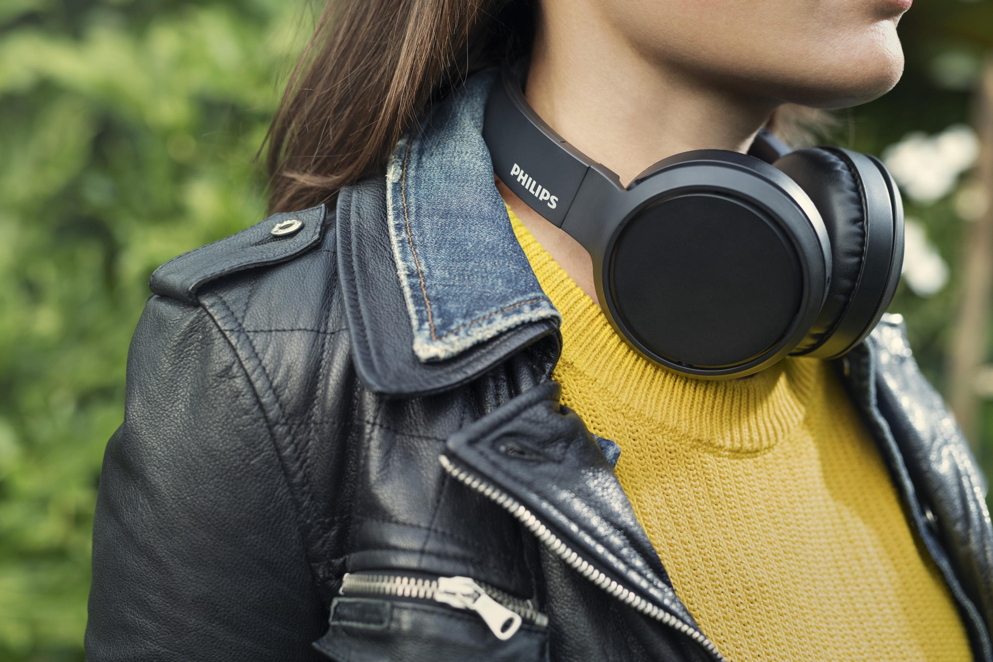 Kobieta z czarnymi słuchawkami na szyi, w czarnej kurtce i żółtym swetrze. Rozmyte zielone tło.