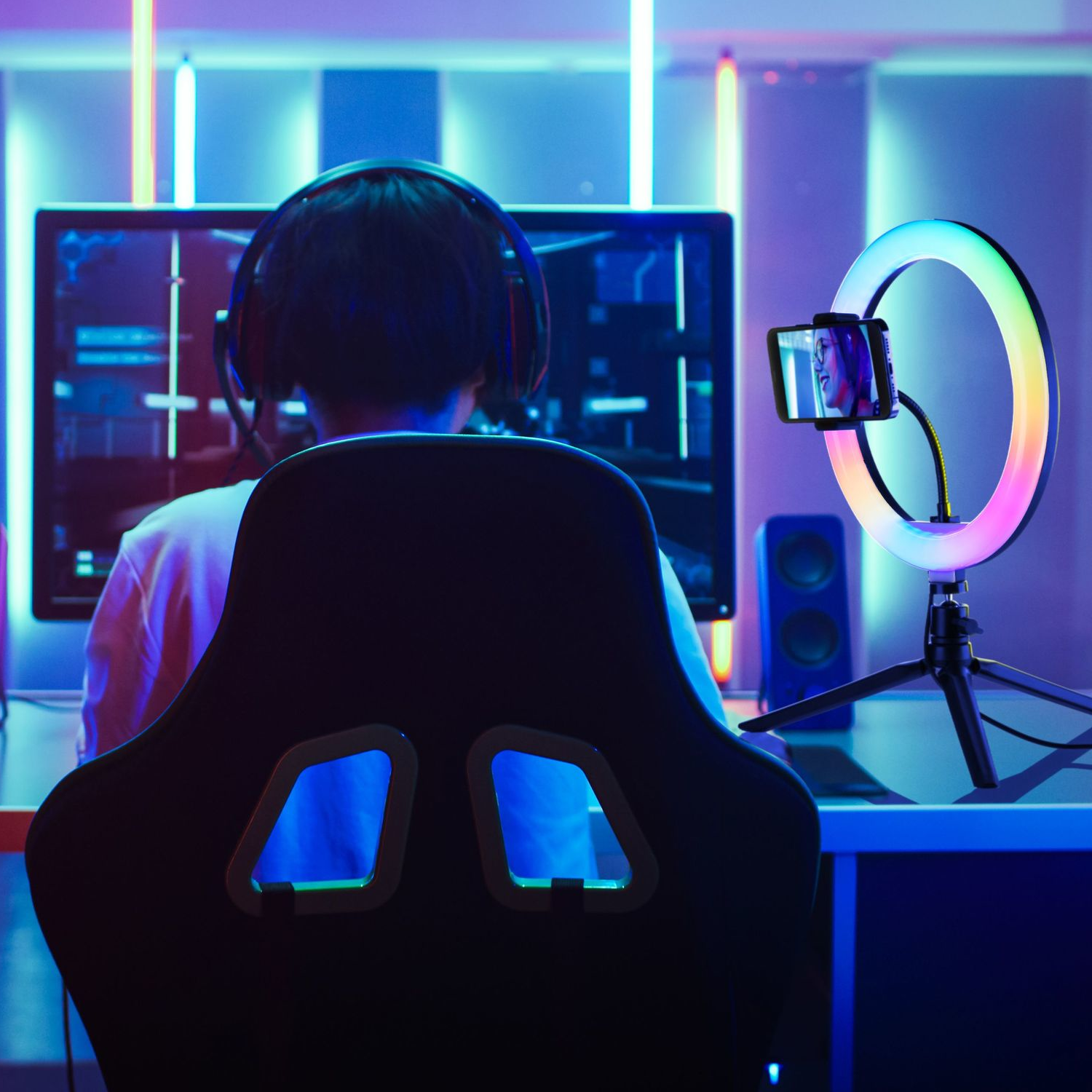 Persona en silla gamer con auriculares, transmitiendo en un monitor y luz de anillo.