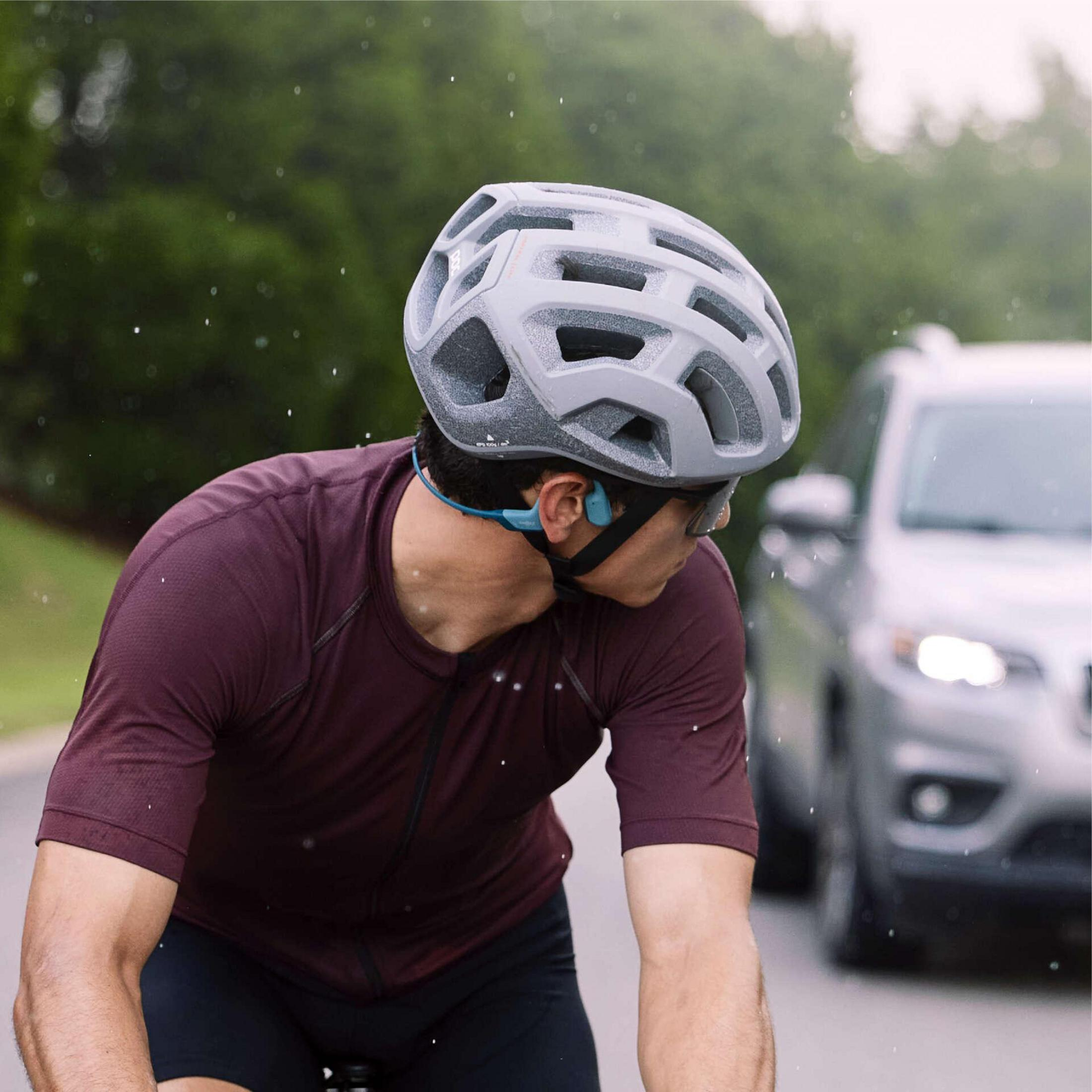 Un ciclista con un casco grigio percorre una strada. Un'auto argentata sullo sfondo.