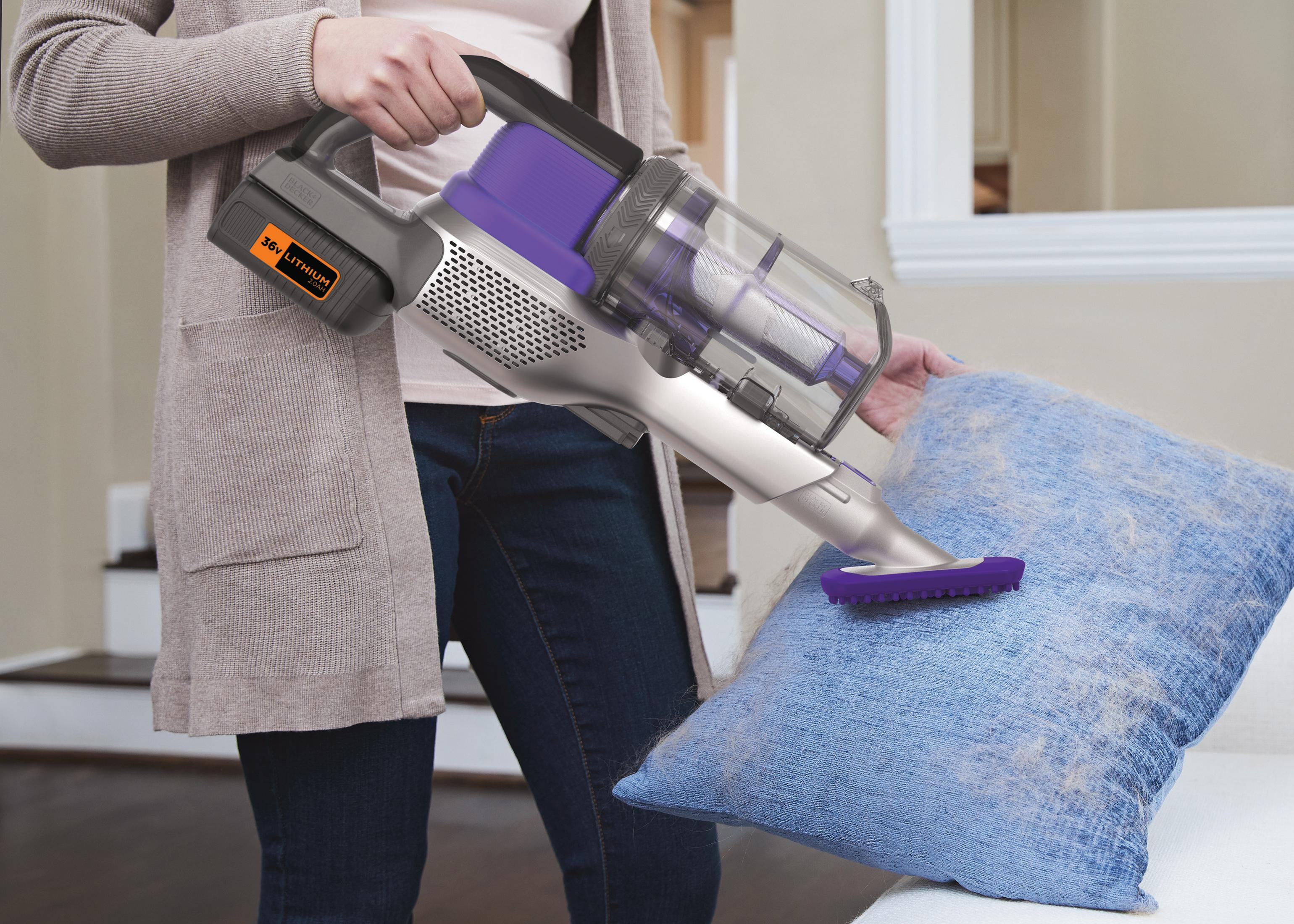 A person vacuums a blue pillow with a gray and purple handheld vacuum cleaner.