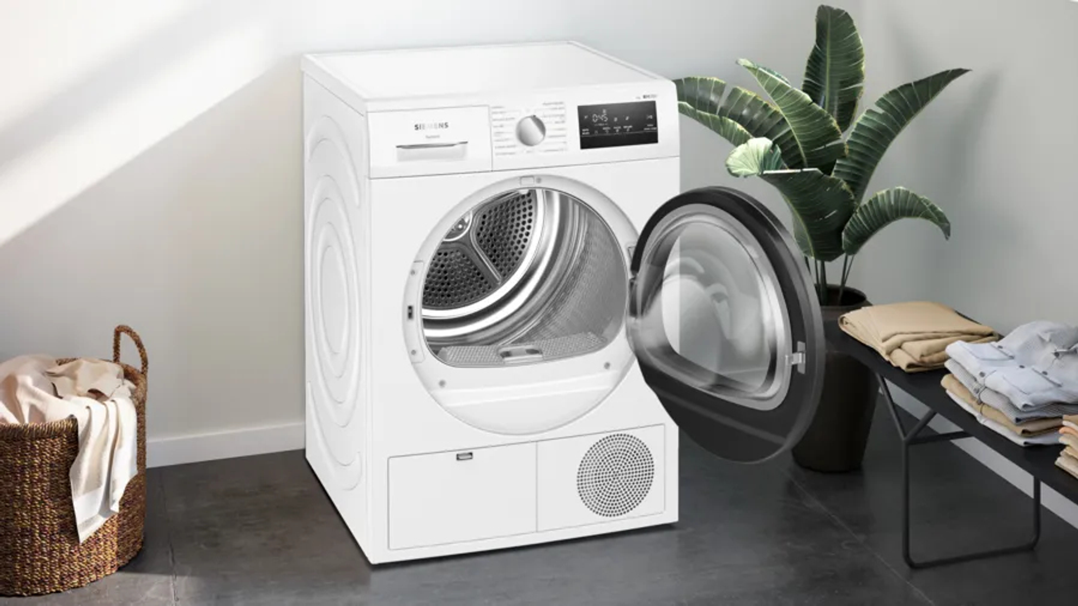Secadora Siemens blanca con puerta abierta. Cesta de ropa y planta detrás.