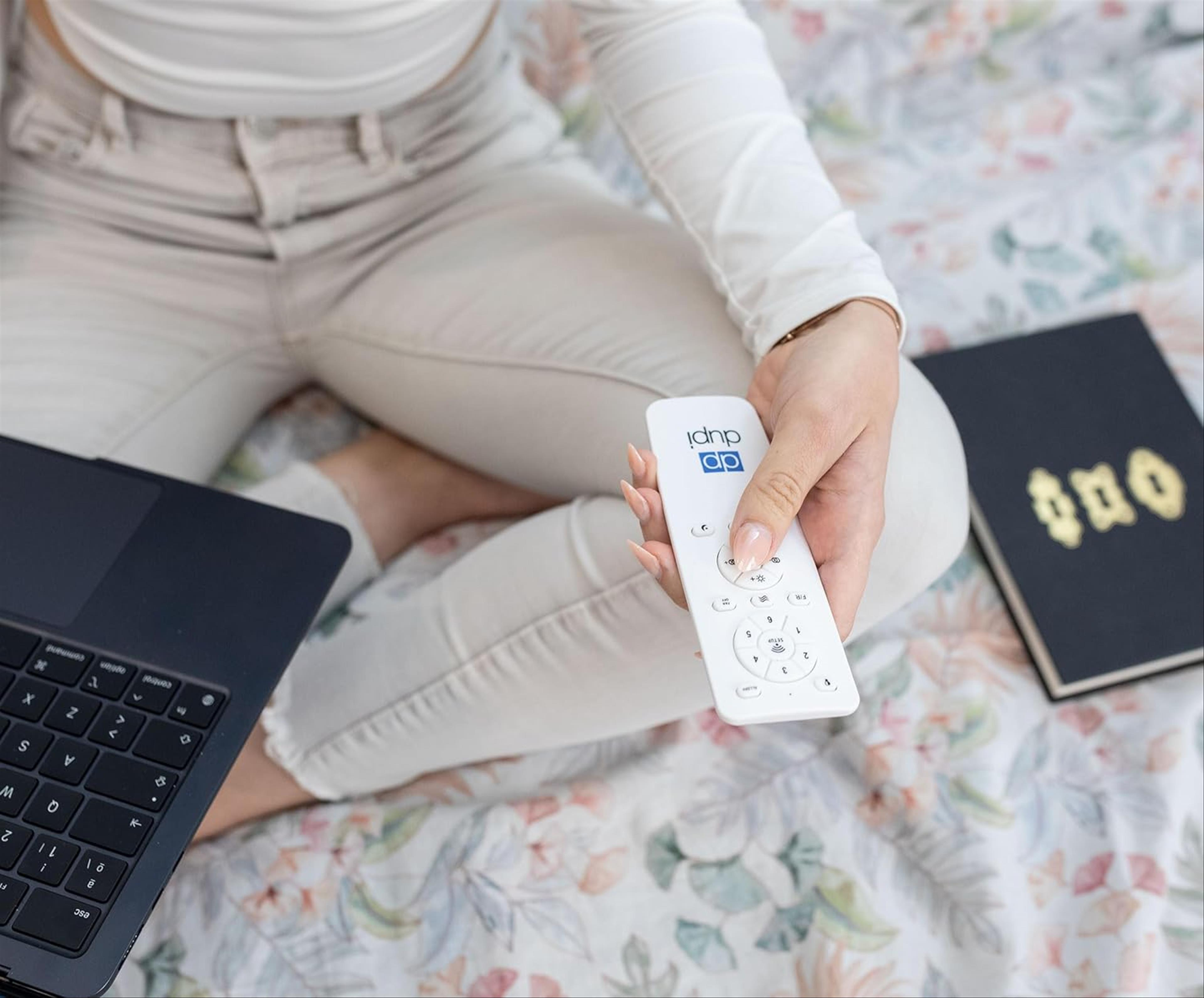 Persona sentada en la cama sosteniendo un control remoto blanco cerca de una laptop y un cuaderno.