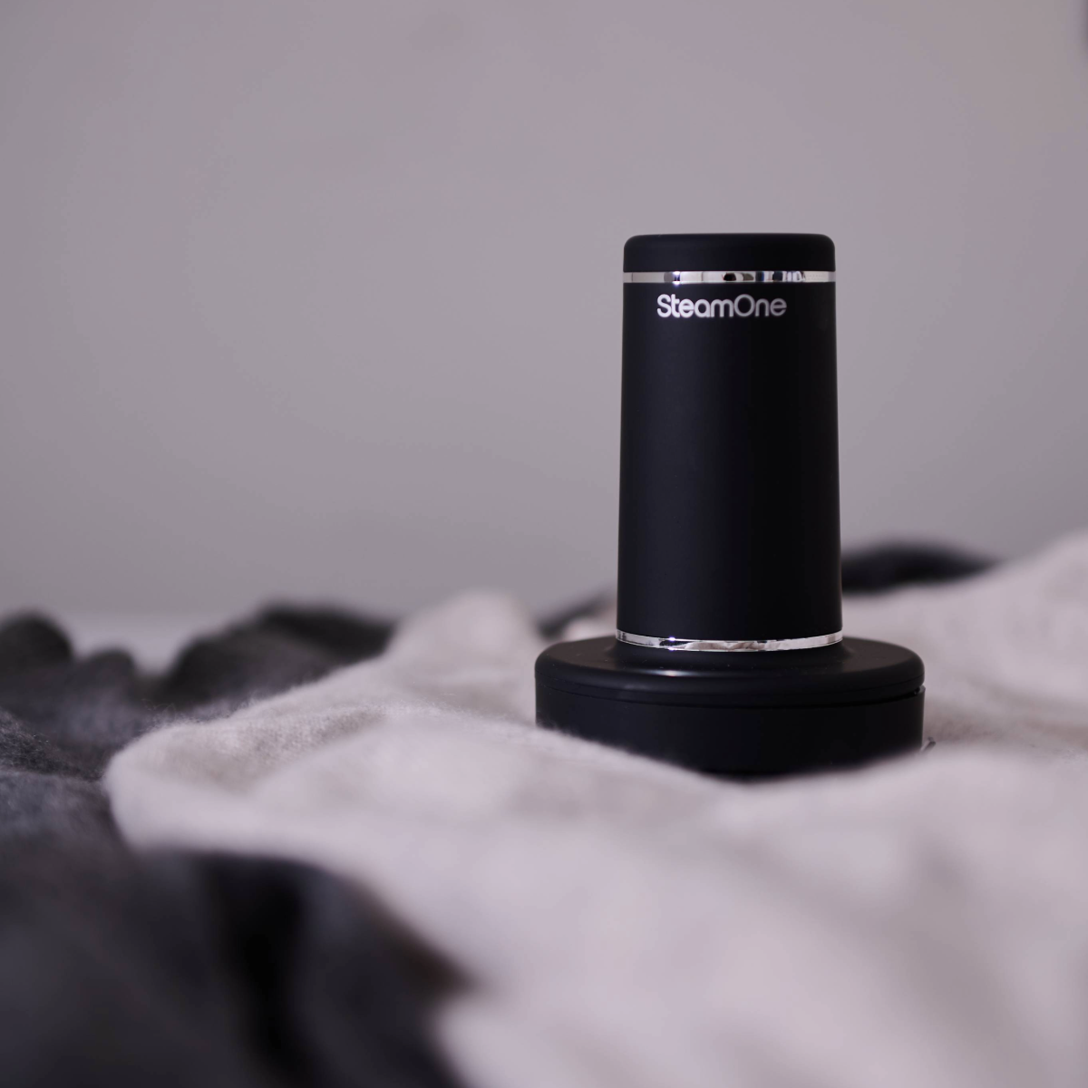 Un petit défroisseur vapeur SteamOne noir est posé sur une couverture blanche. L'arrière-plan est gris flou.