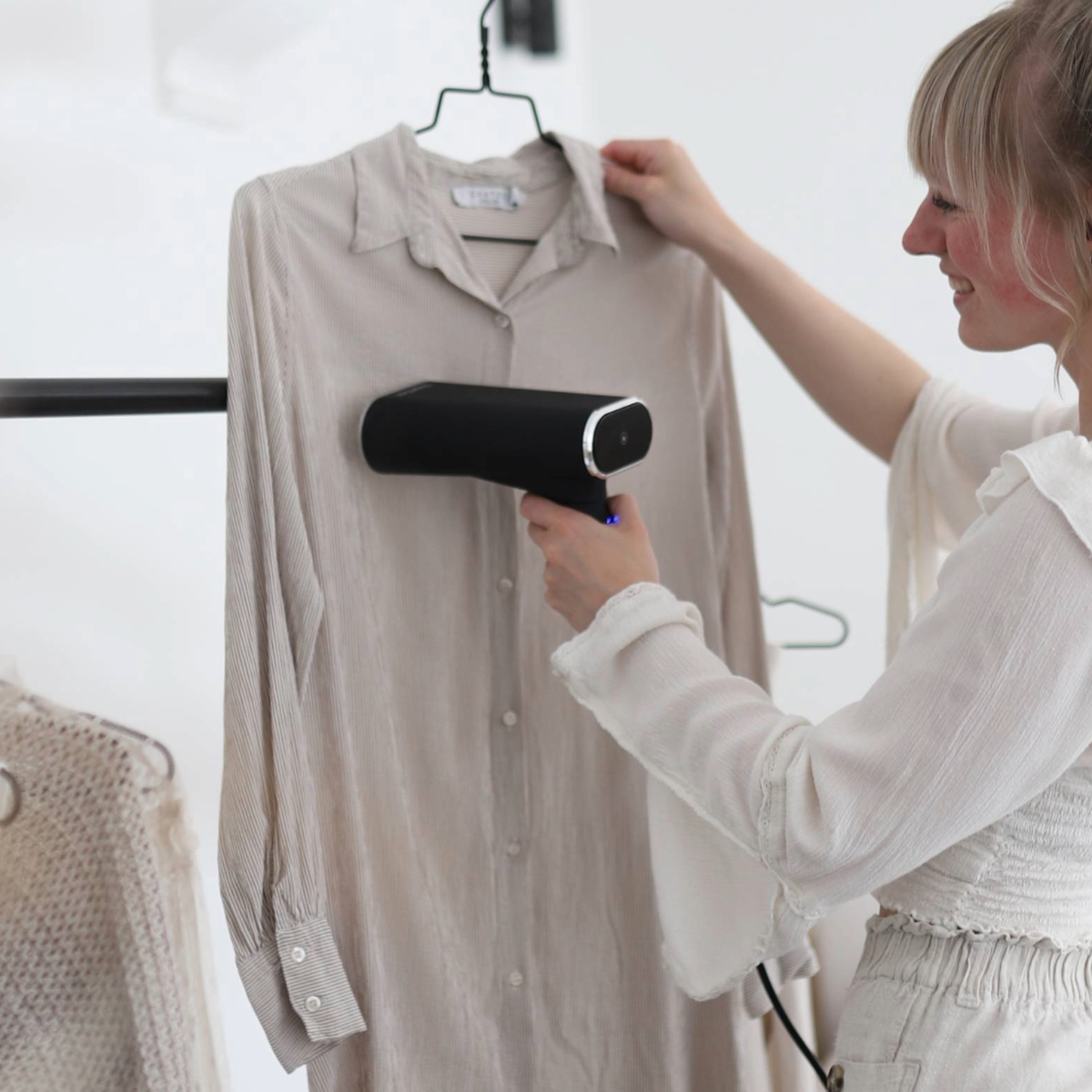 Une femme défroisse une chemise avec un défroisseur à main. D'autres vêtements sont sur un portant en arrière-plan.