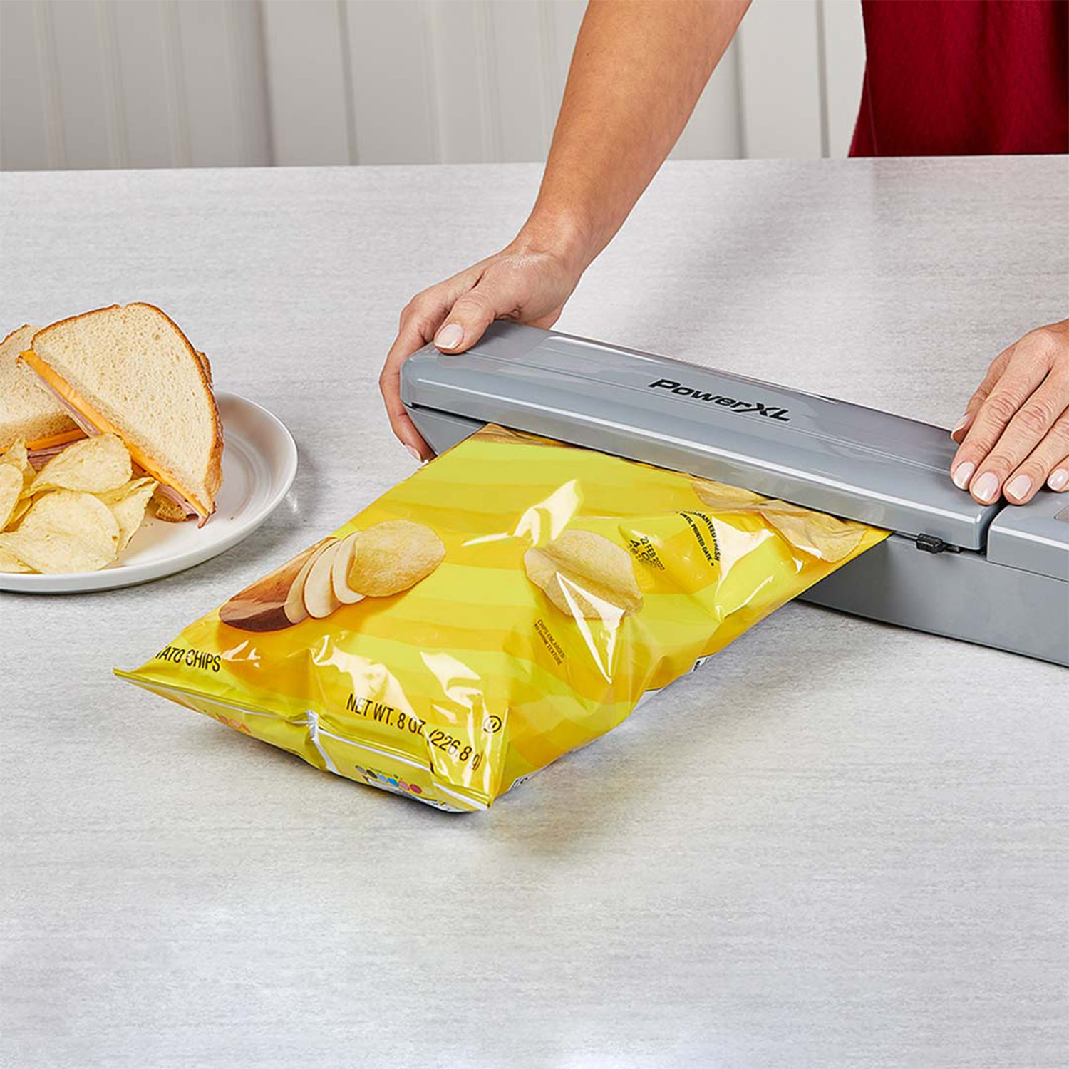 Hände versiegeln einen gelben Kartoffelchip-Beutel mit einem Siegelgerät, Teller mit Chips und Sandwich.