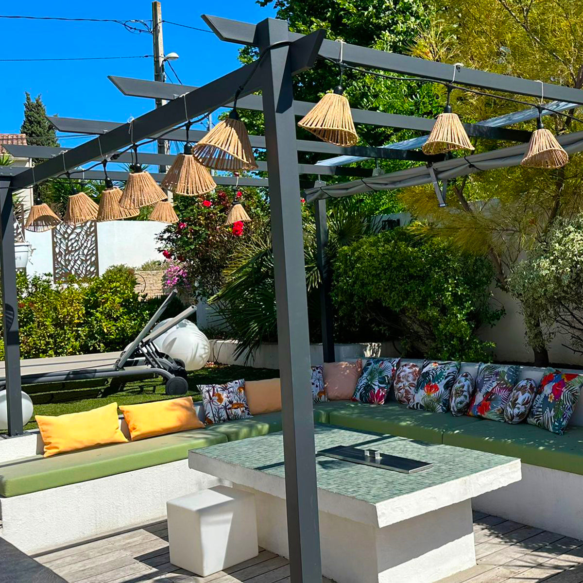 Terrasse mit Pergola, Sitzgelegenheiten und Tisch. Dekorative Lampen hängen von der Pergola, Pflanzen ringsherum.
