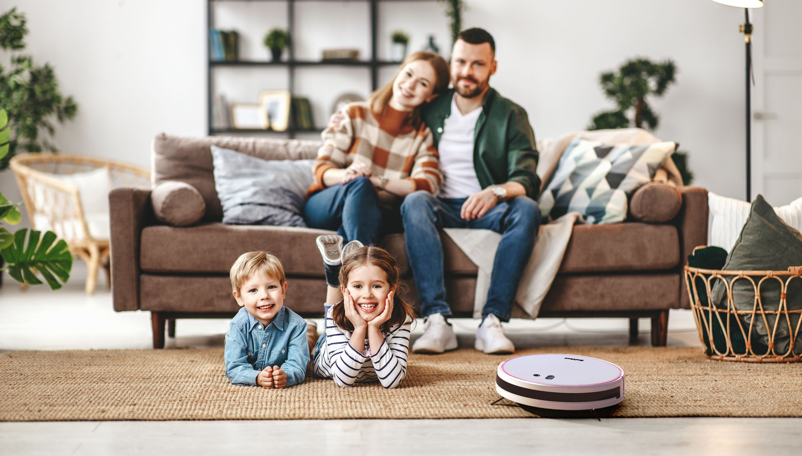 Vierköpfige Familie lächelt auf einem Teppich mit einem Roboterstaubsauger. Ein Sofa ist im Hintergrund.