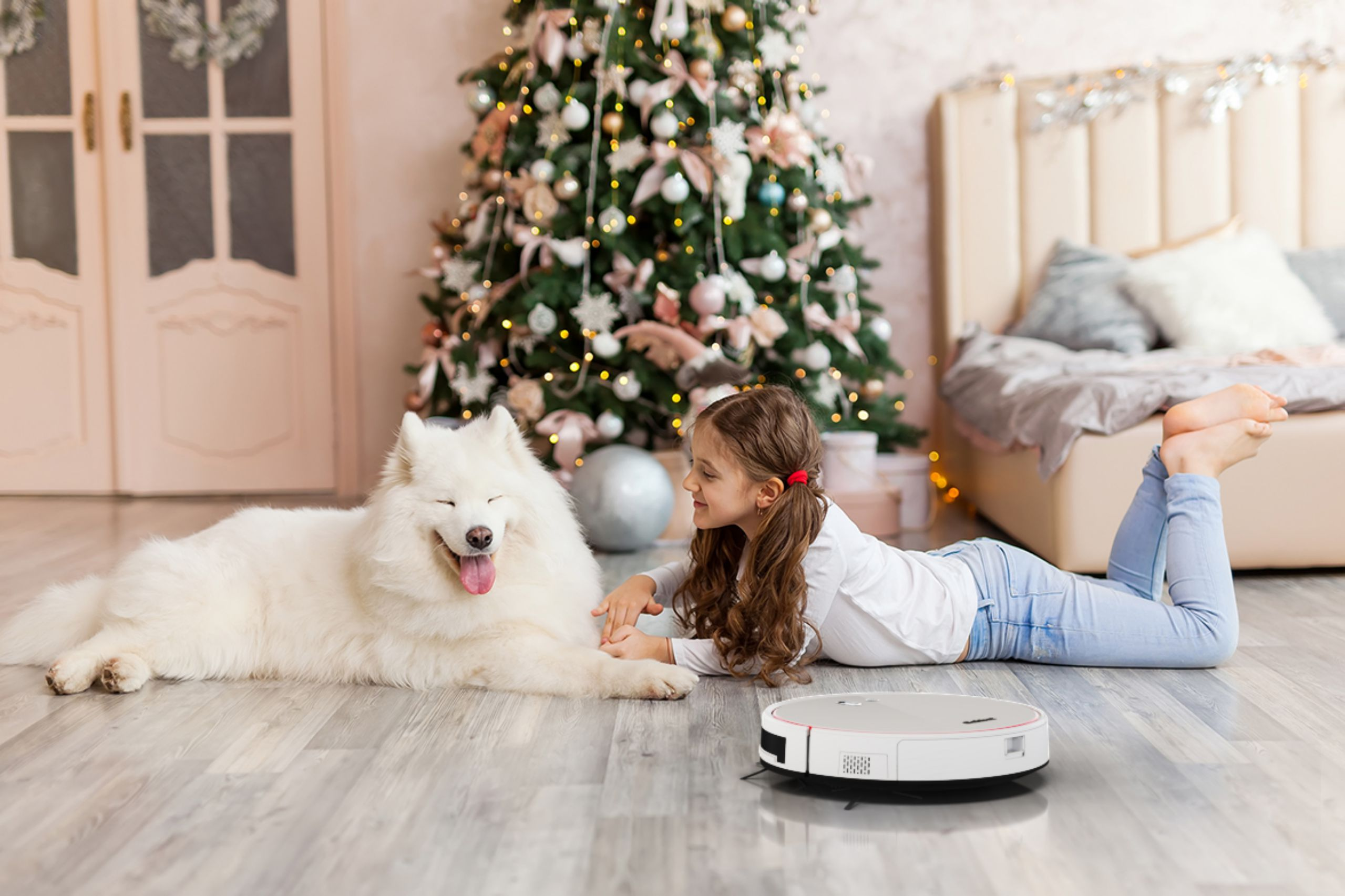 Mädchen und Hund liegen am Boden in der Nähe eines Weihnachtsbaums und eines Saugroboters.