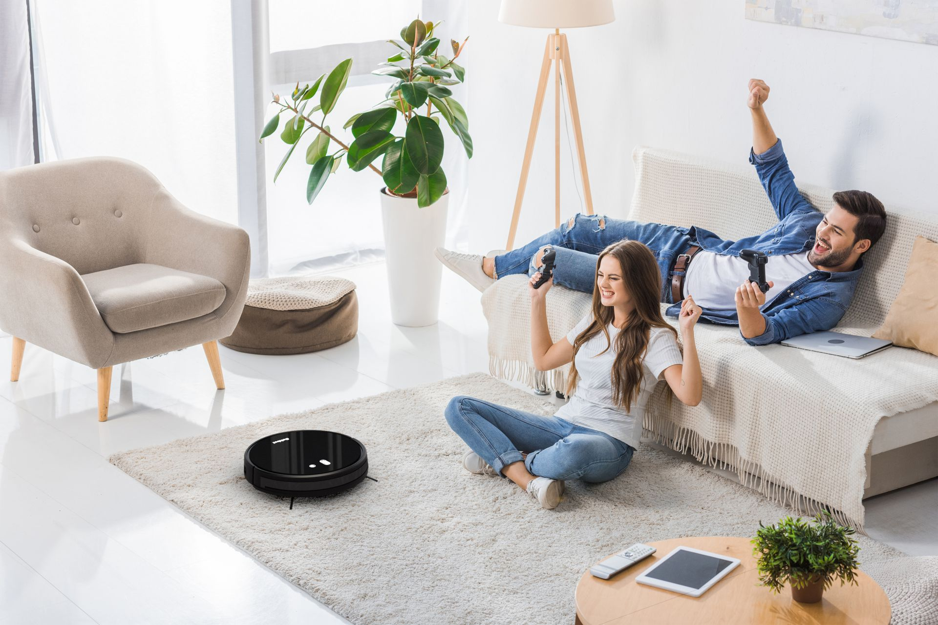 Paar spielt Videospiele auf einem Sofa, ein Roboter-Staubsauger auf dem Teppich.