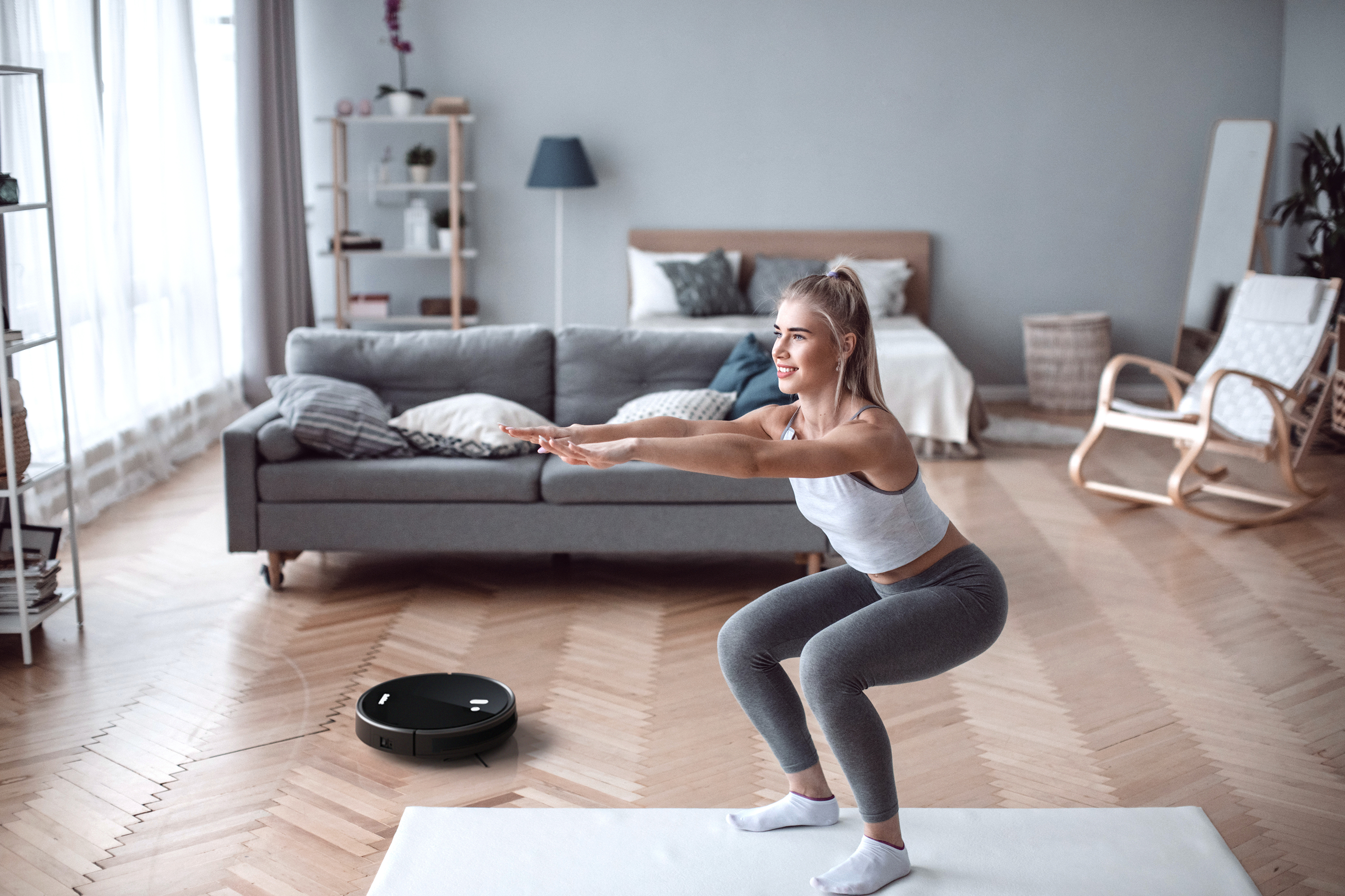 Frau macht Kniebeugen im Wohnzimmer. Ein Roboter-Staubsauger steht auf dem Boden. Graues Sofa und Bett im Hintergrund.