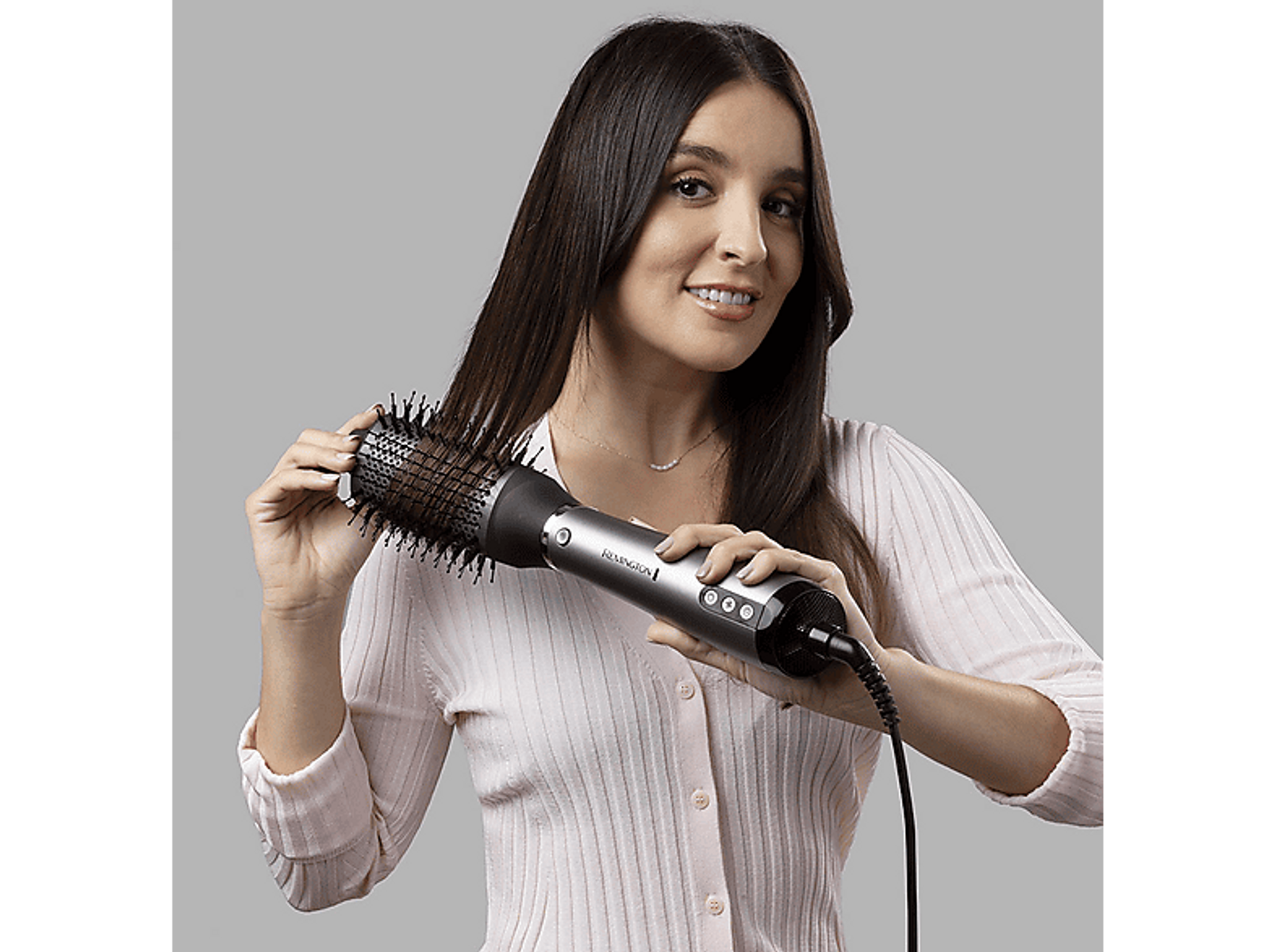 Donna con i capelli castani che usa uno styler per capelli. Sorride mentre si sistema i capelli su sfondo grigio.