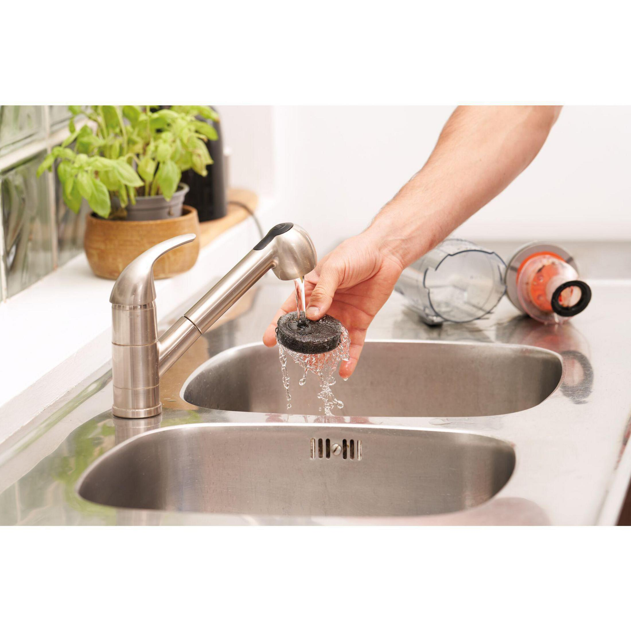 Una mano che risciacqua un filtro nero sotto un rubinetto da cucina cromato in un lavandino doppio.