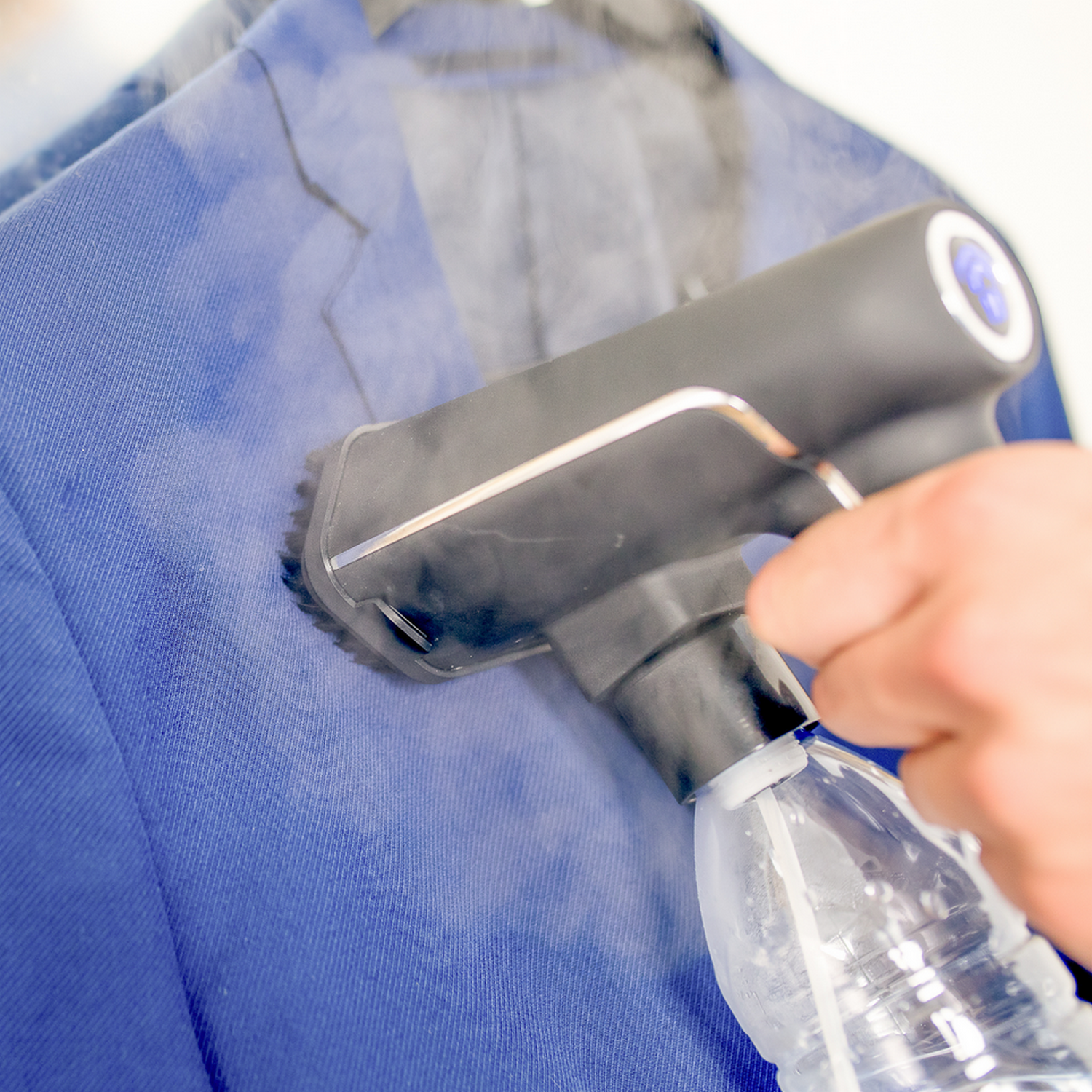 Eine Hand benutzt ein Dampfbügeleisen auf einer blauen Jacke, mit einer Wasserflasche.