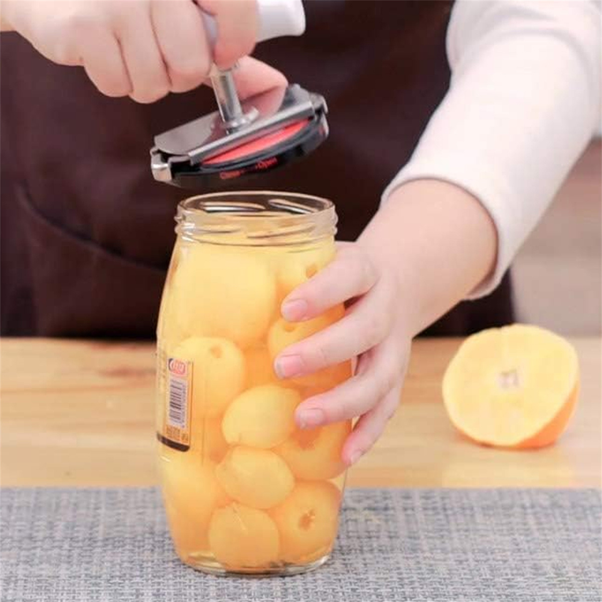 Person benutzt einen Gläseröffner für ein Glas mit gelben Früchten. Orange daneben.